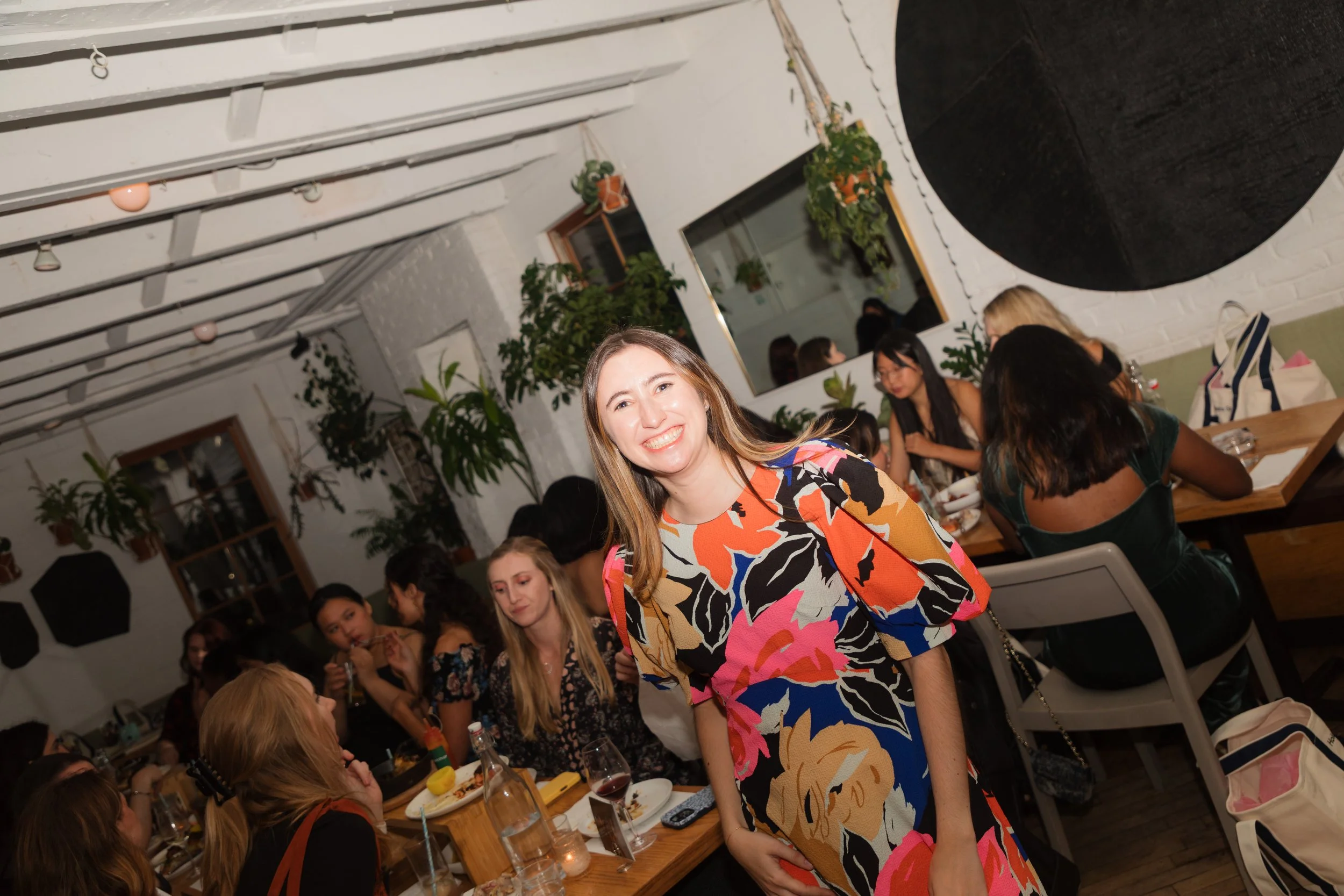A woman in a colorful floral dress smiling at the camera at a dinner gathering with other women seated at a wooden table in a cozy, decorated restaurant with plants and white brick walls.