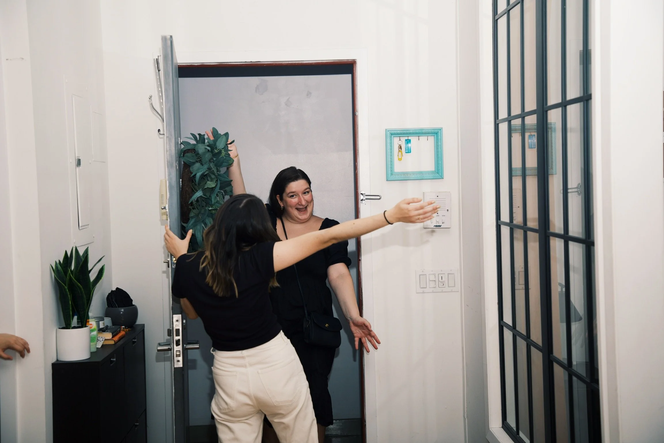 Two women greet each other at the doorway of a home, smiling and gesturing enthusiastically with open arms.