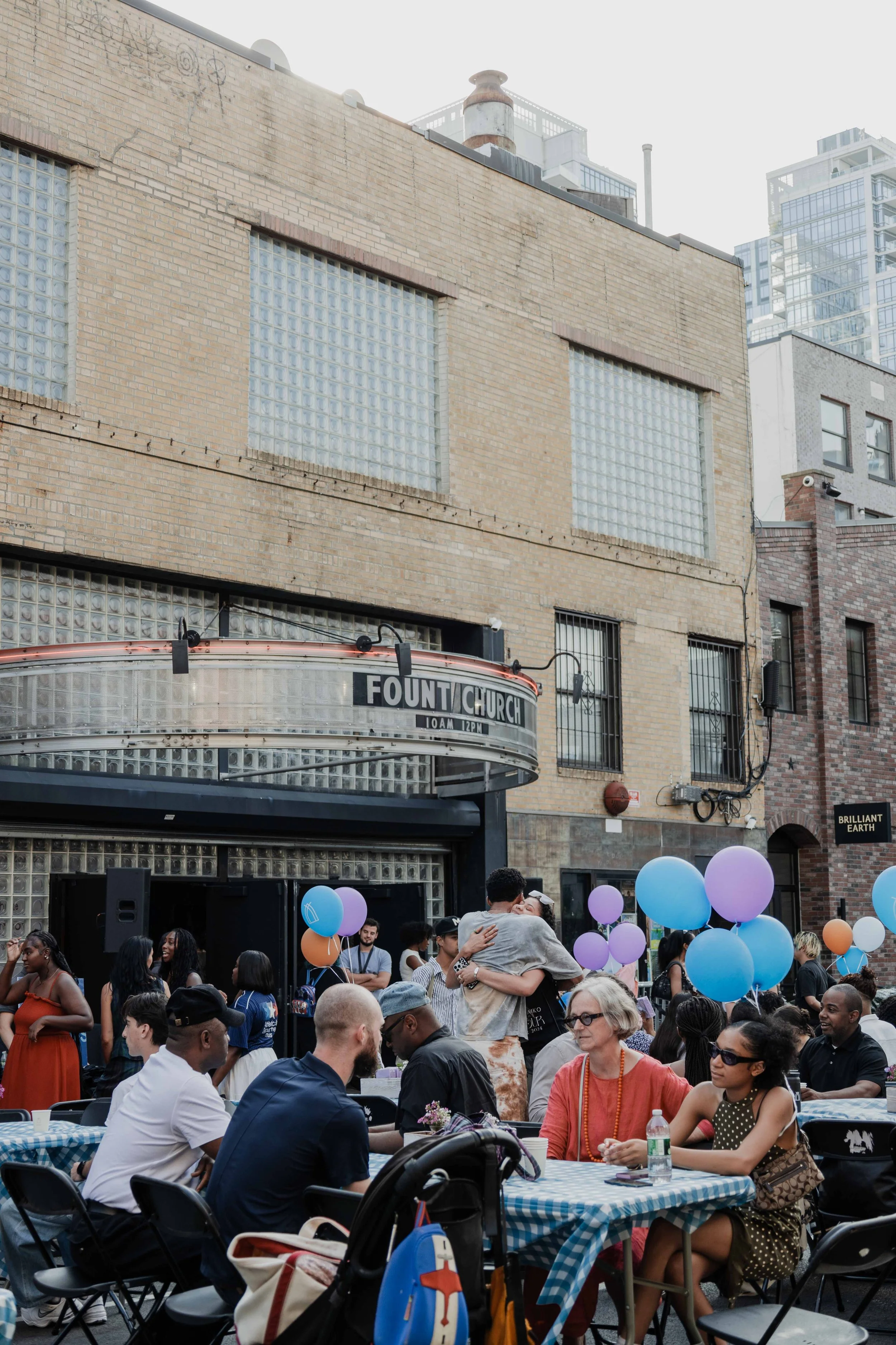 People gathered outdoors at a community event with balloons, sitting at checkered tables, and embracing in front of a building with a marquee reading 'Fount Church'.