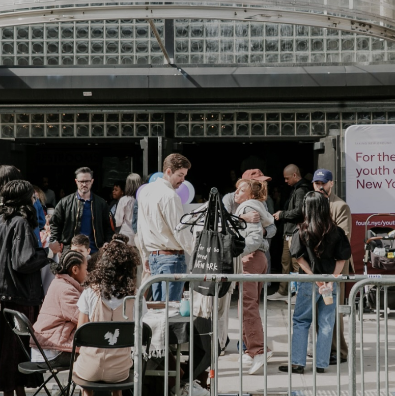 People gathered outdoors at an event, some sitting and others standing, with a man holding a baby and a woman hugging nearby, in front of a building with glass blocks and signs.