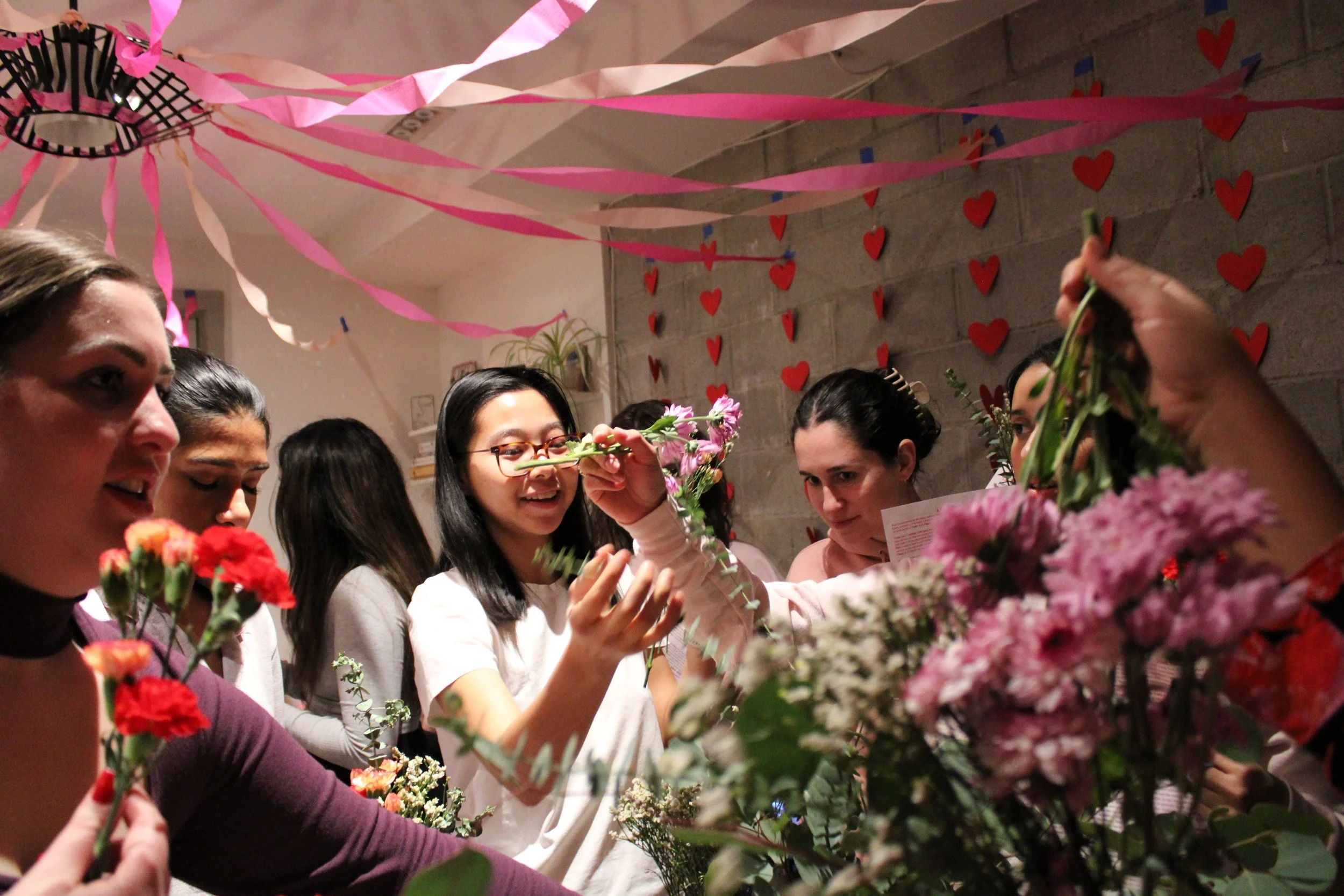Group of women celebrating Valentine's Day with flowers, pink and red heart decorations, and pink streamers hanging from the ceiling.