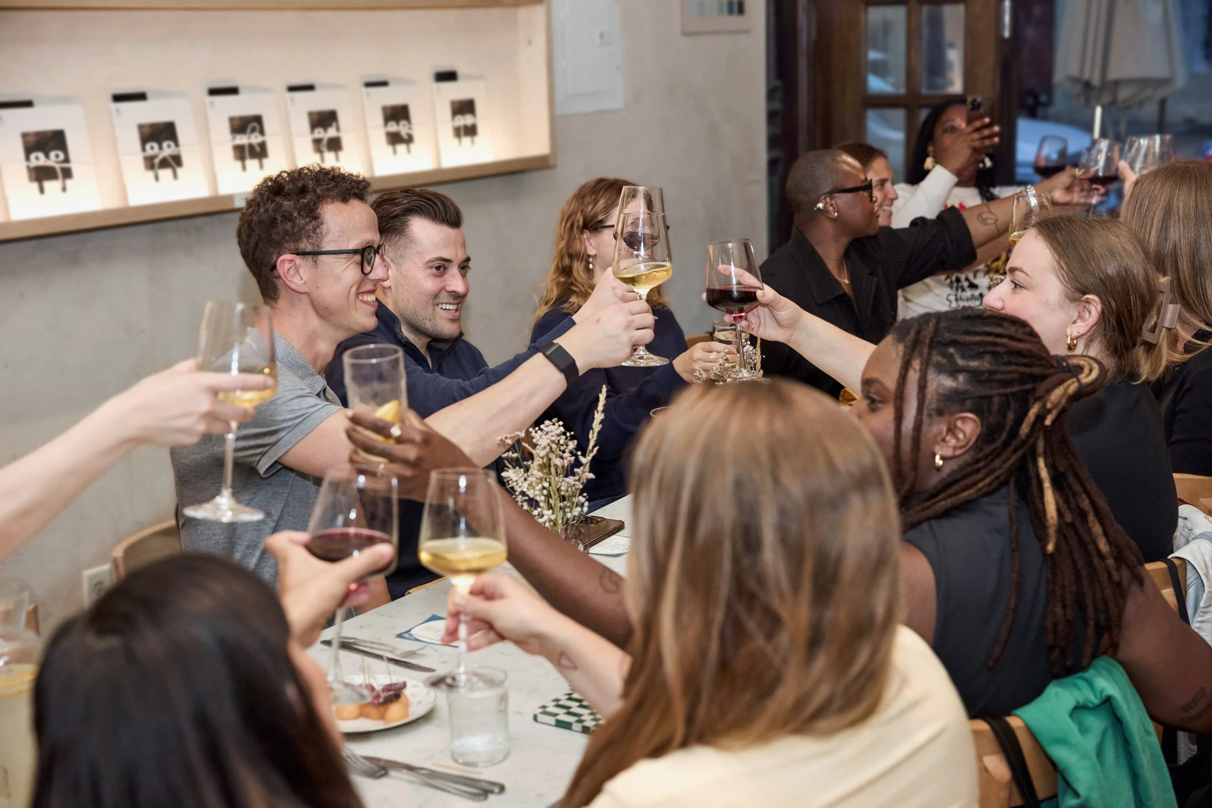 A group of diverse people at a dinner party raising glasses of wine in a toast, smiling and celebrating together in a cozy restaurant setting.