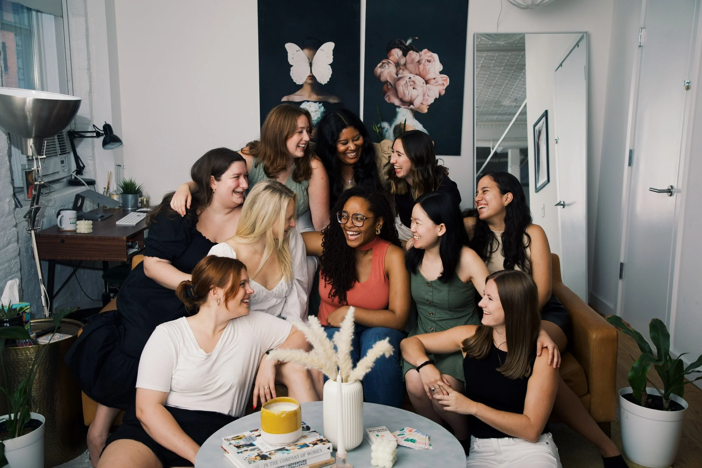 A group of women gathered in a cozy living room, smiling and laughing together, with some sitting on a sofa and others standing behind them.