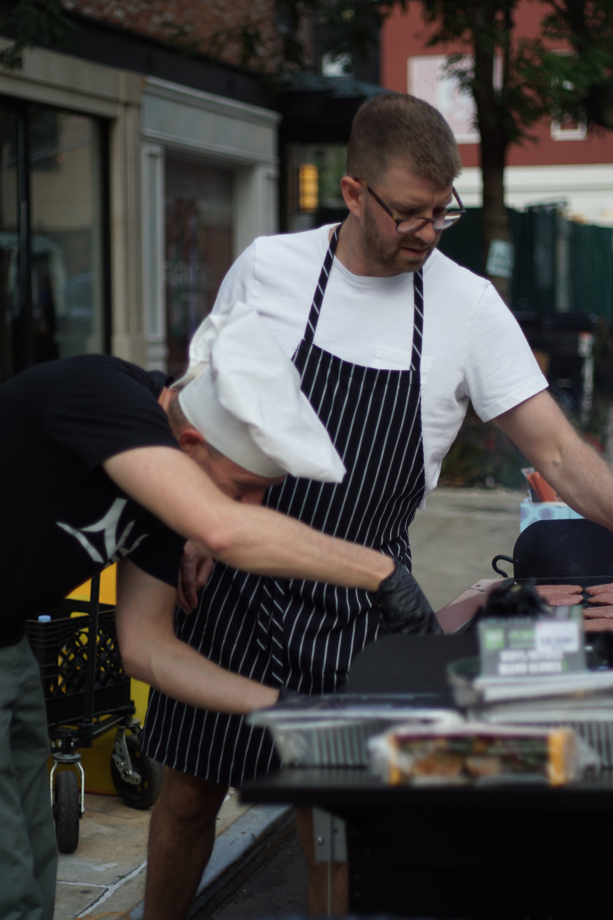 Two men cooking hamburgers outdoors, one wearing a chef's hat and black T-shirt, and the other wearing glasses, a white T-shirt, and a striped apron.