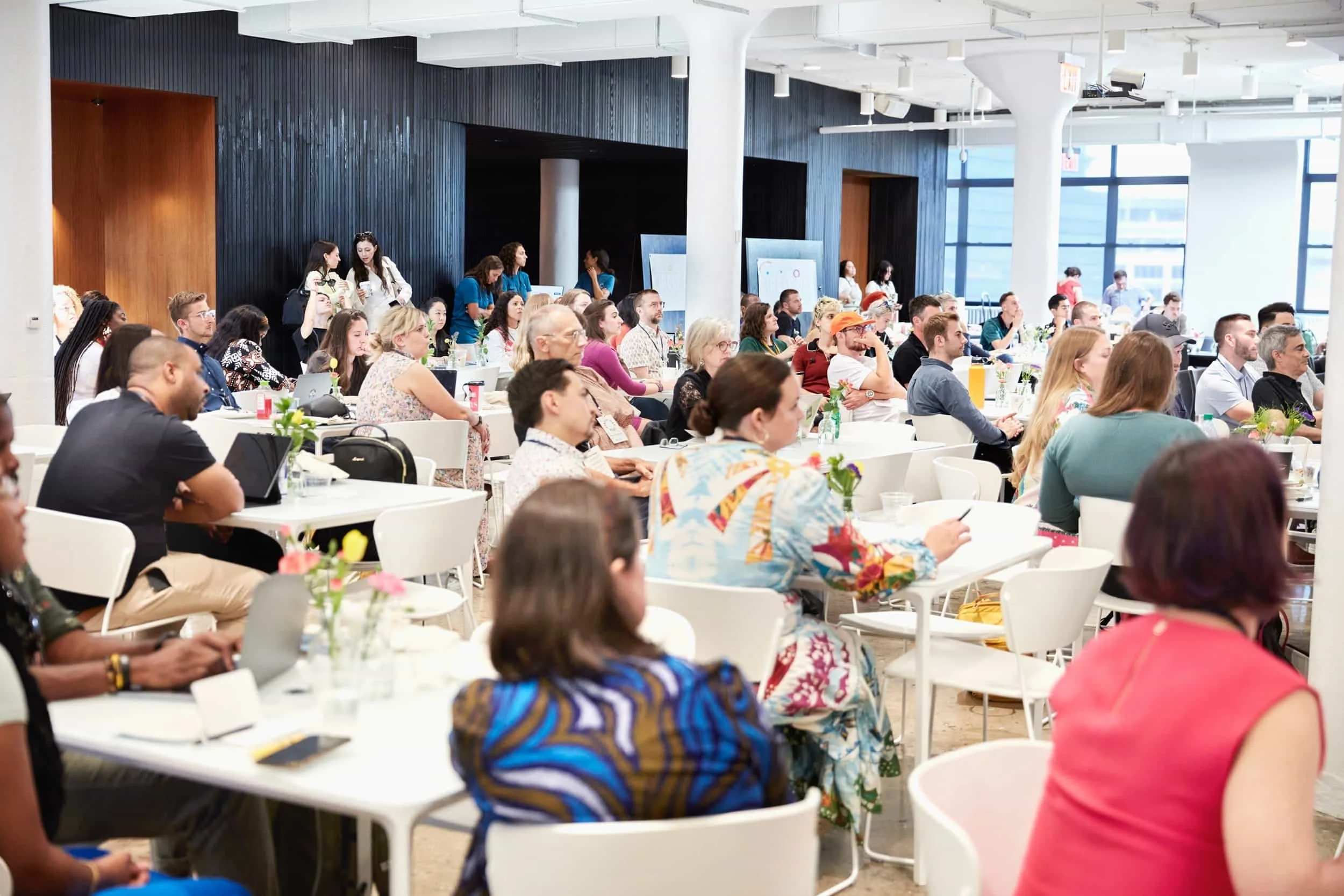 A diverse group of people seated at tables in a bright conference room, attentively watching a conference presentation.