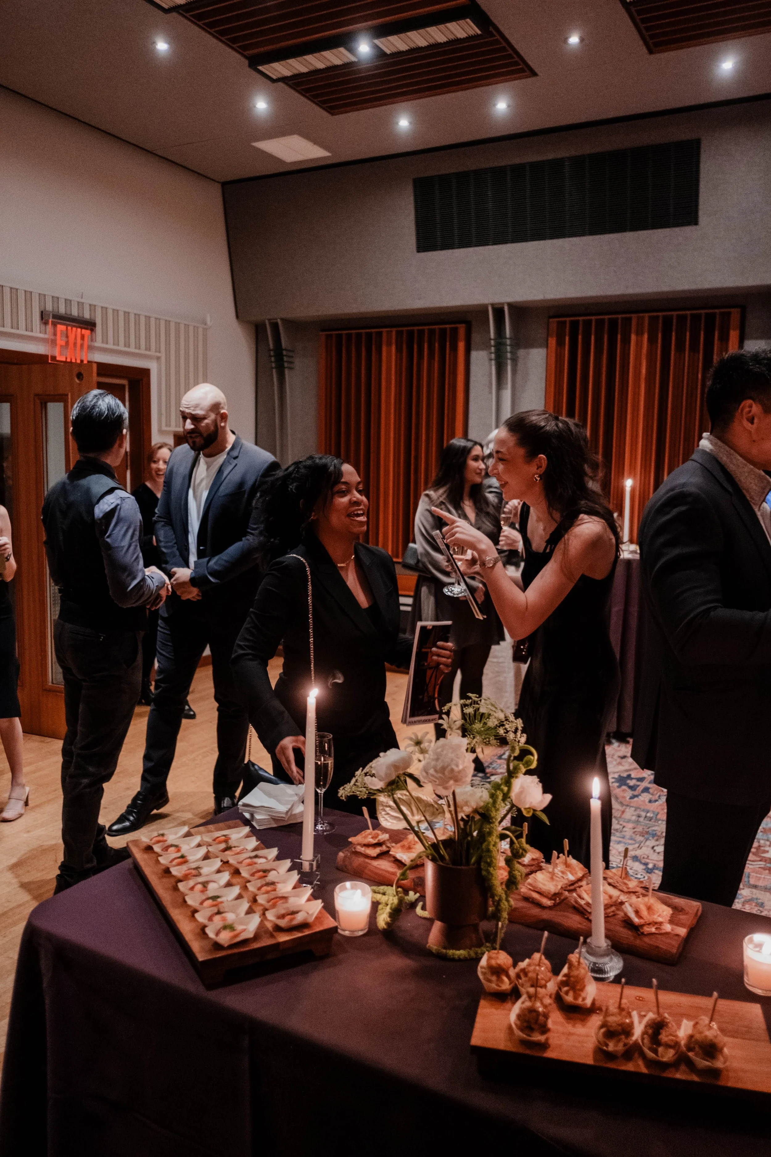 People socializing at an indoor event with a table of appetizers, flowers, and lit candles.
