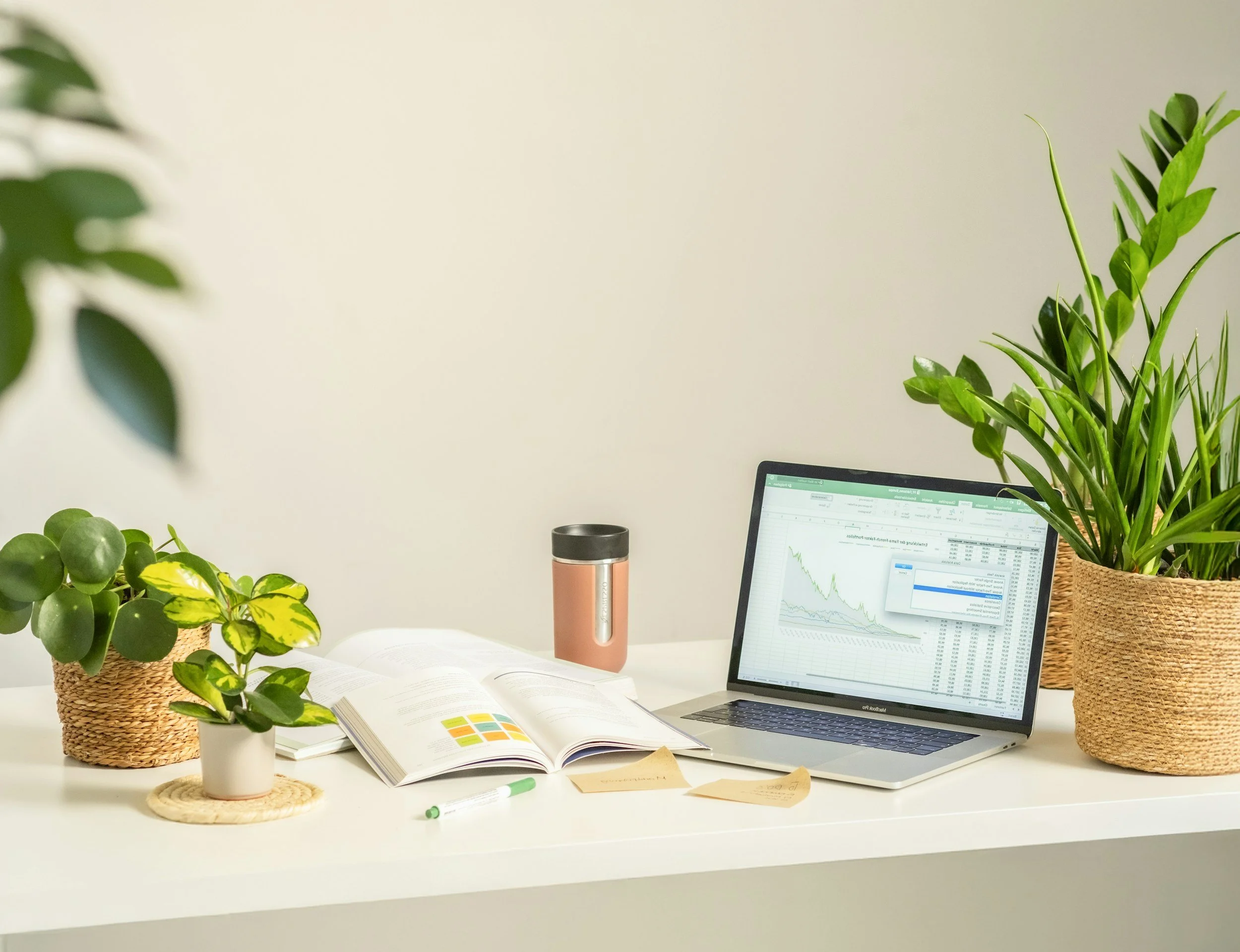 A tidy workspace featuring a white desk decorated with potted houseplants, an open book, a pen, a laptop displaying financial charts, a stainless steel tumbler, and sticky notes. The background has a plain white wall.
