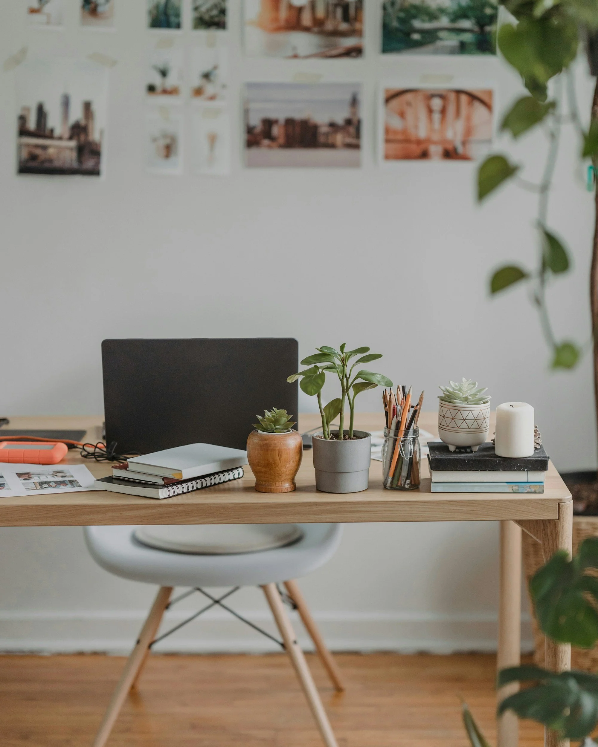 A workspace with a wooden table holding potted plants, notebooks, a glass jar with pencils, candles, and a laptop. In the background, there is a white wall with photographs, a black chair, and a white dining chair with wooden legs.
