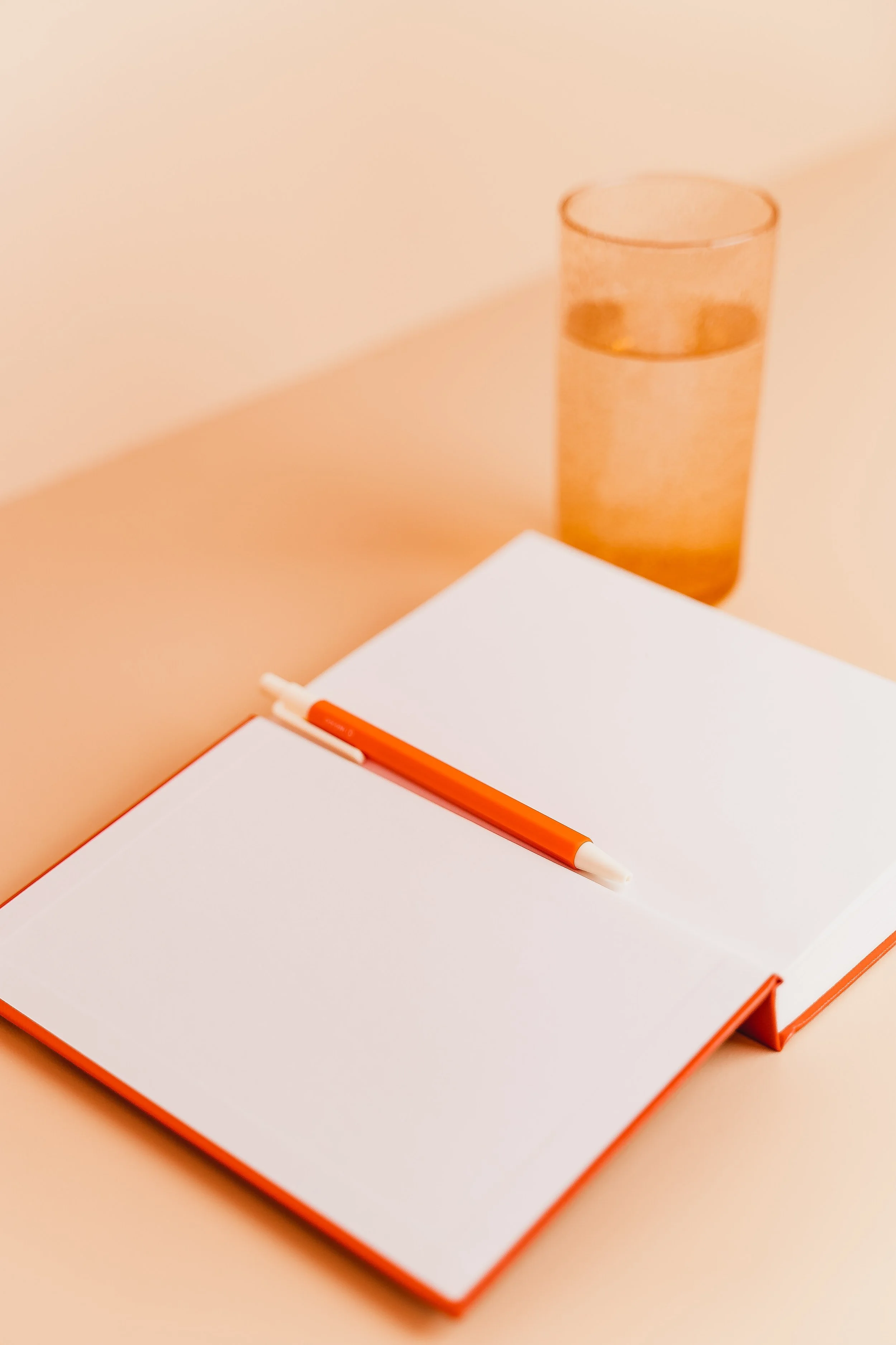 Open notebook with blank white pages, orange pen on top, and a tall glass of water on a peach-colored surface and background.