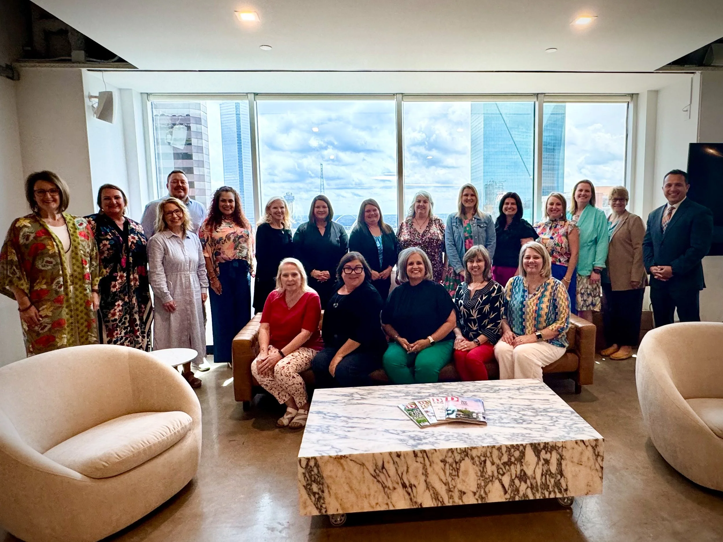 Group of 20 people in a modern indoor space with large windows and city skyline view, sitting and standing around a sofa and marble coffee table.