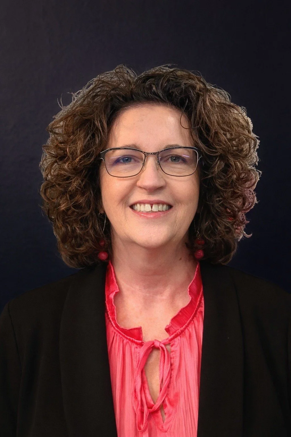 A woman with dark brown hair, glasses, pearl earrings, and a pearl necklace, wearing a black blazer and a pink top, smiling in front of a dark blue background.