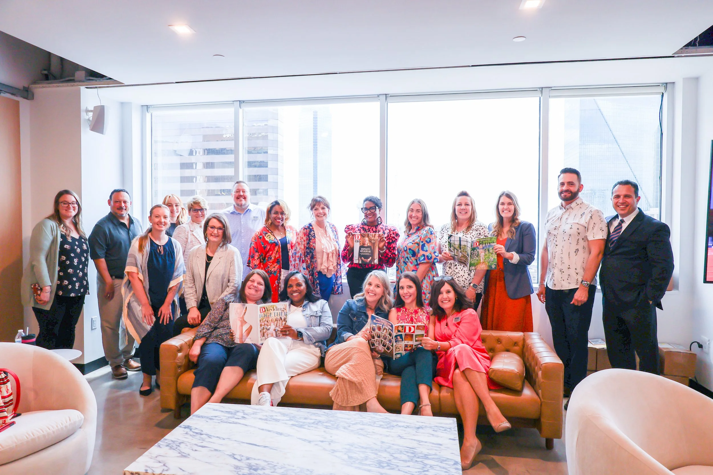 Group of women and men posing together in a modern office lounge with large windows and cityscape views, some sitting on a sofa and others standing behind.