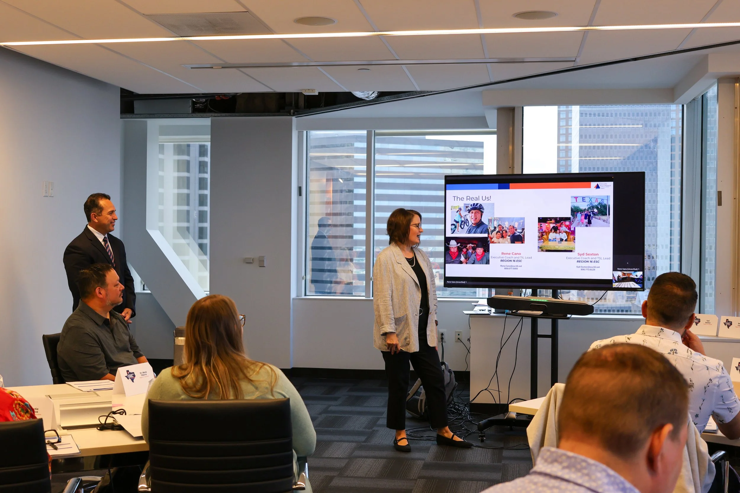 A woman presenting in a conference room with a large screen displaying a presentation. Several attendees are seated and taking notes.