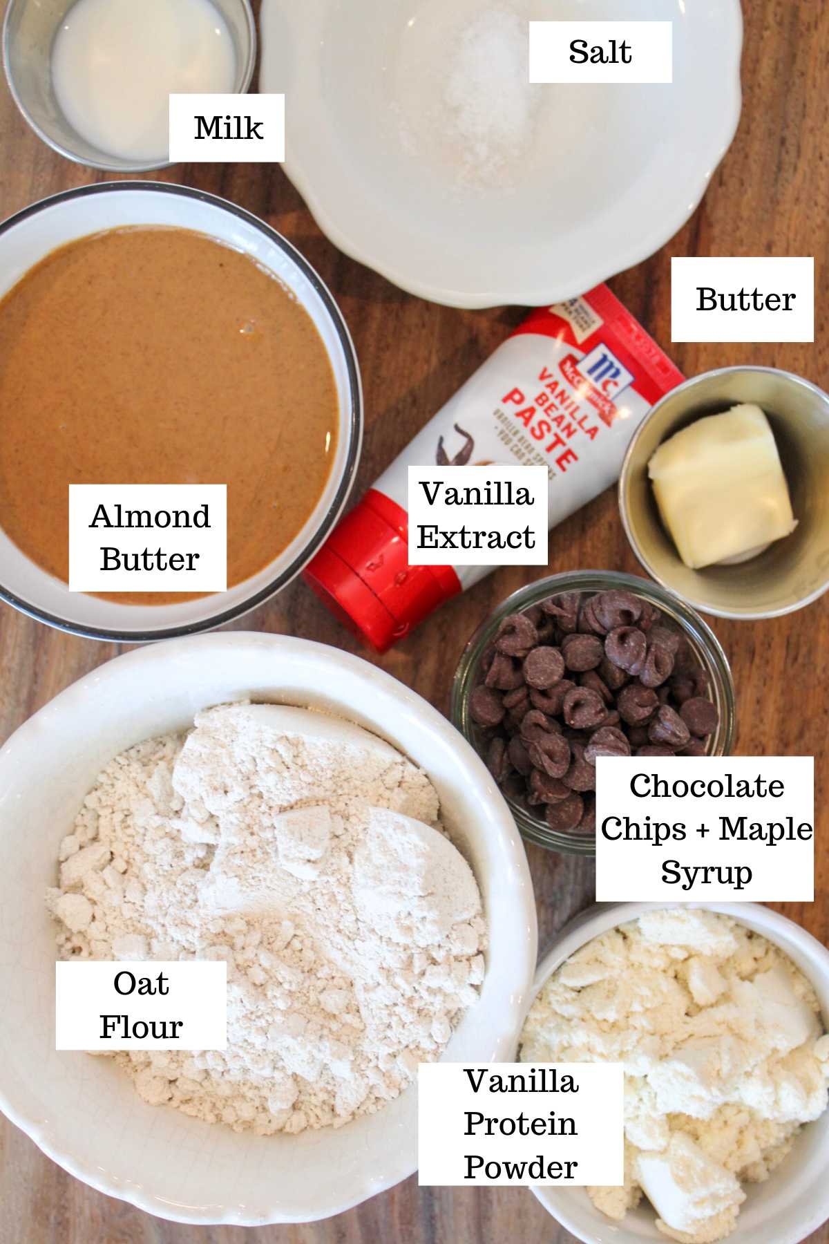 A top view of baking ingredients labeled on a wooden surface: almond butter, oat flour, vanilla protein powder, chocolate chips, milk, salt, butter, and vanilla extract.