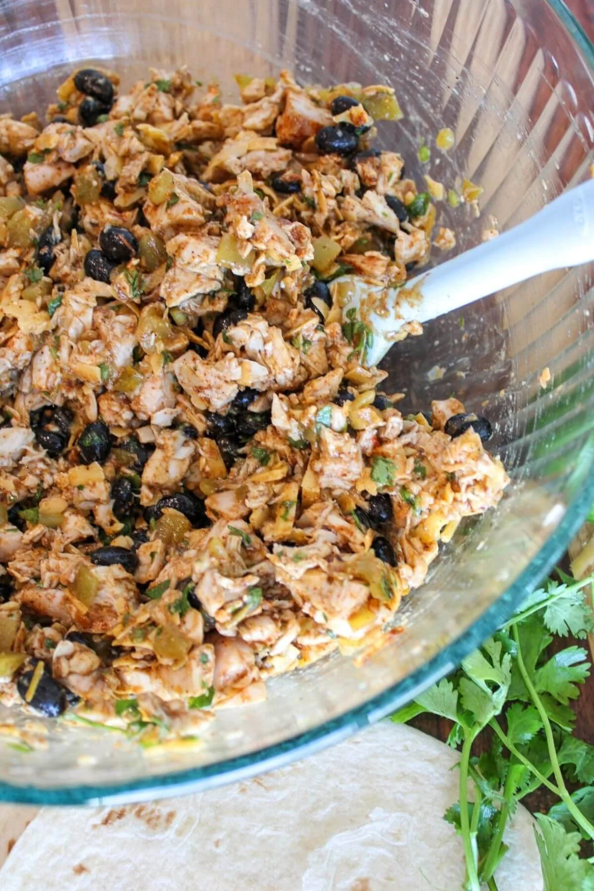 Overhead view of a savory shredded chicken mixture with black beans, green chiles, and herbs in a glass bowl beside fresh cilantro.