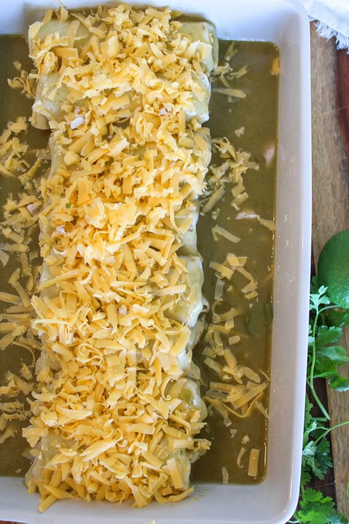 Baked enchiladas covered in melted shredded cheese and green sauce fill a white casserole dish, with fresh cilantro and lime nearby.