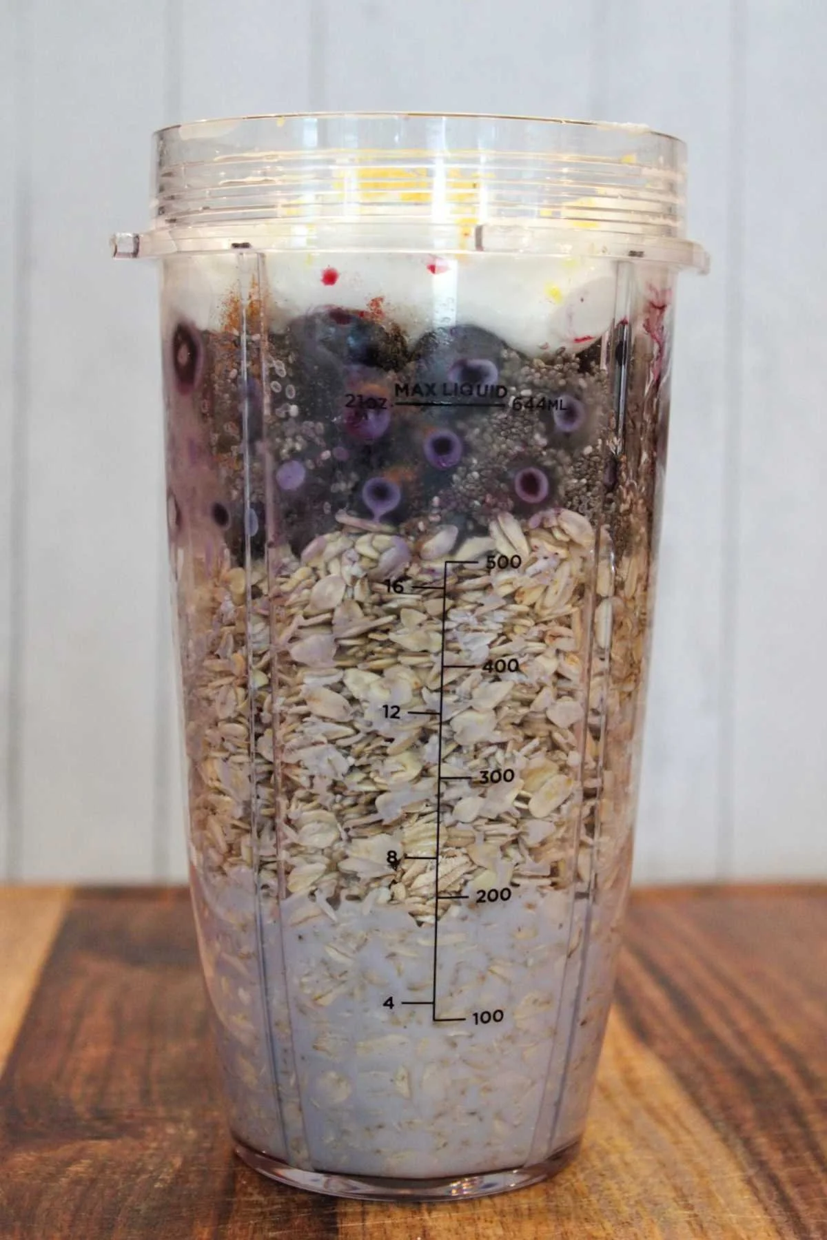 A clear blender cup filled with layers of milk, oats, blueberries, chia seeds, and yogurt sits on a wooden table, ready for blending.