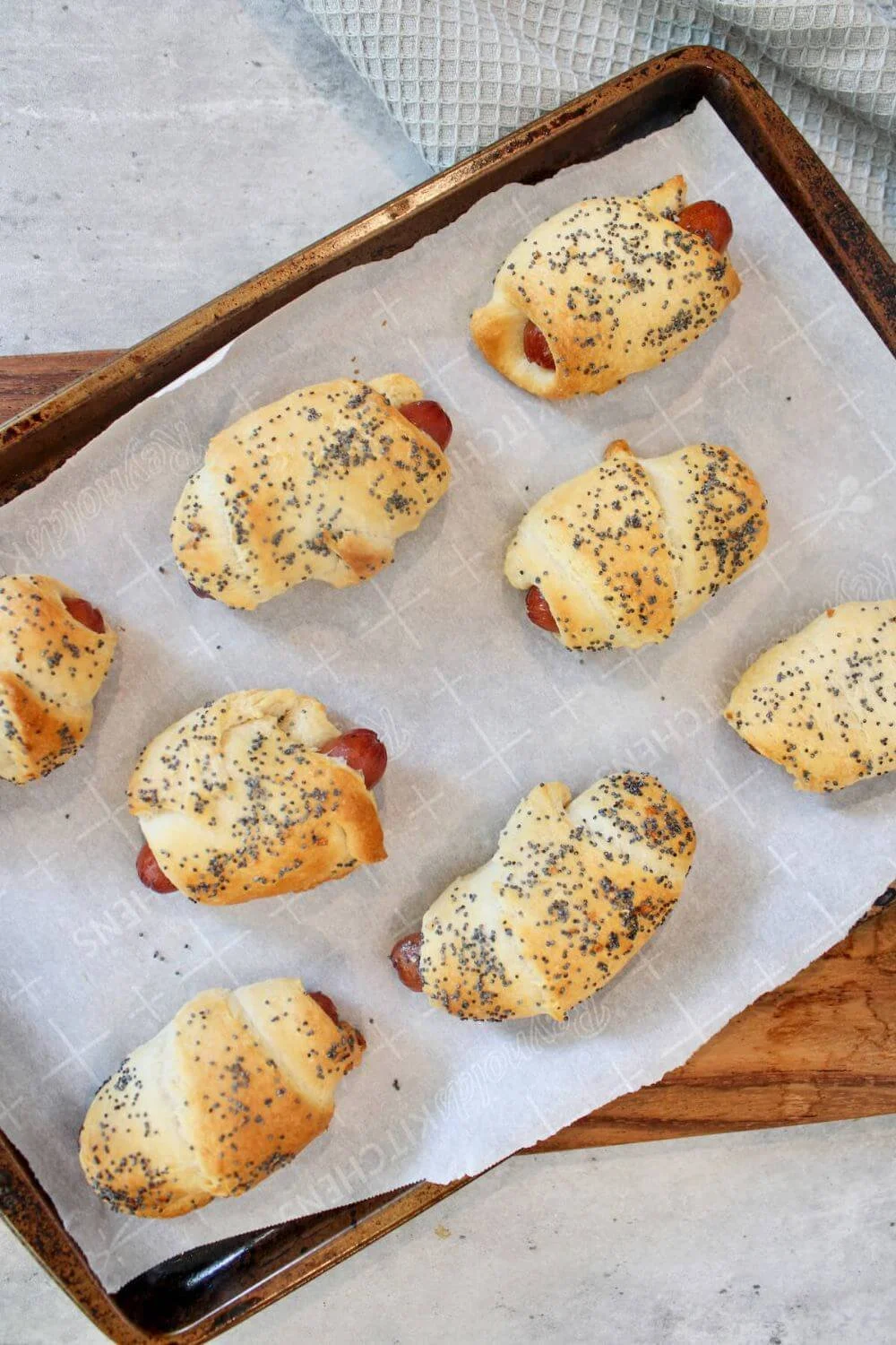 Eight golden-brown lil smokies pigs in a blanket topped with poppy seeds on a parchment-lined baking sheet, set against a light countertop.