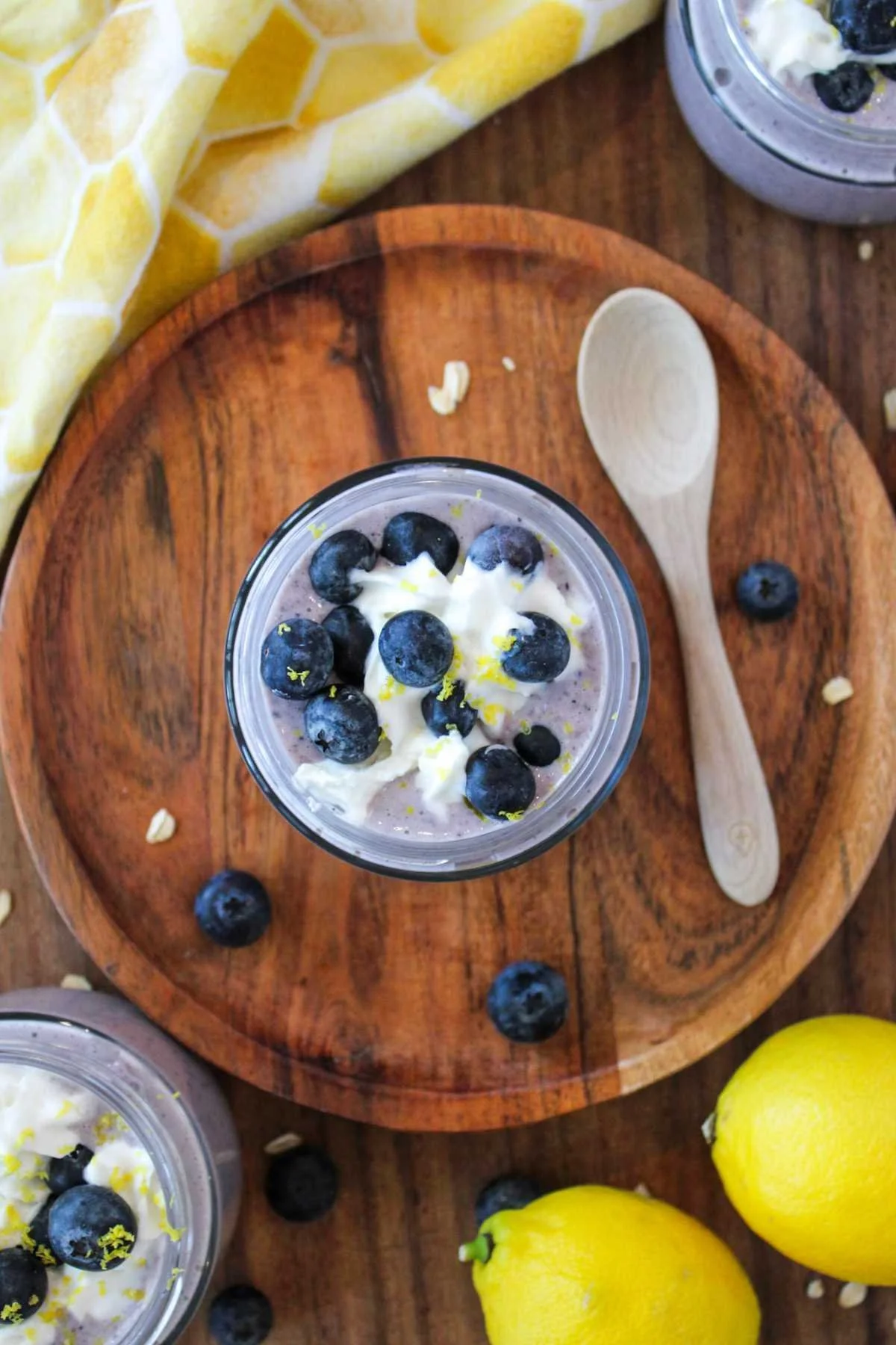A creamy blueberry overnight oats in a jar with fresh blueberries and lemon zest on top, placed on a wooden board with a spoon and lemons nearby.