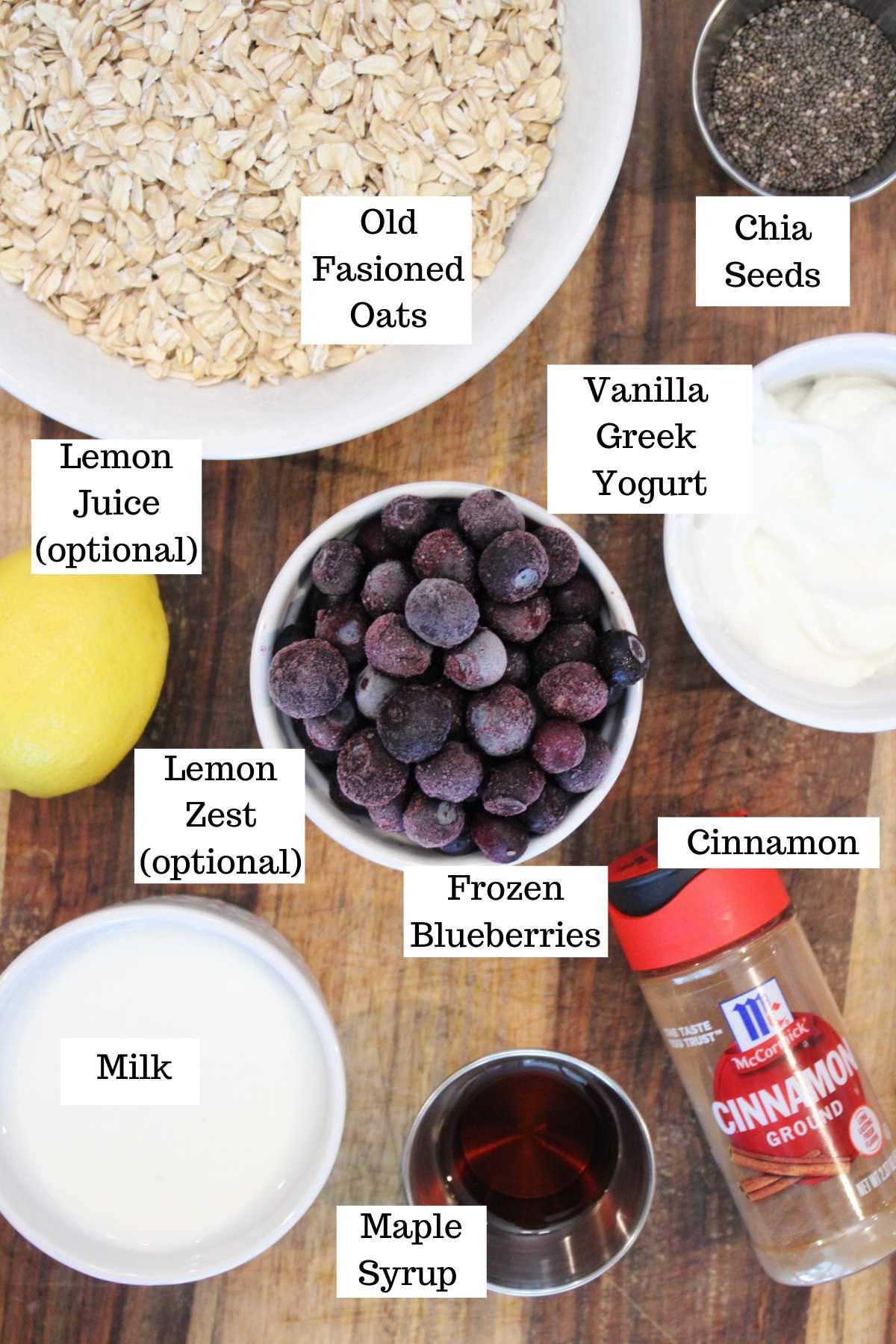 Ingredients for a breakfast recipe are labeled: old fashioned oats, chia seeds, vanilla Greek yogurt, frozen blueberries, milk, maple syrup, cinnamon, lemon, and optional lemon juice and zest, all on a wooden surface.
