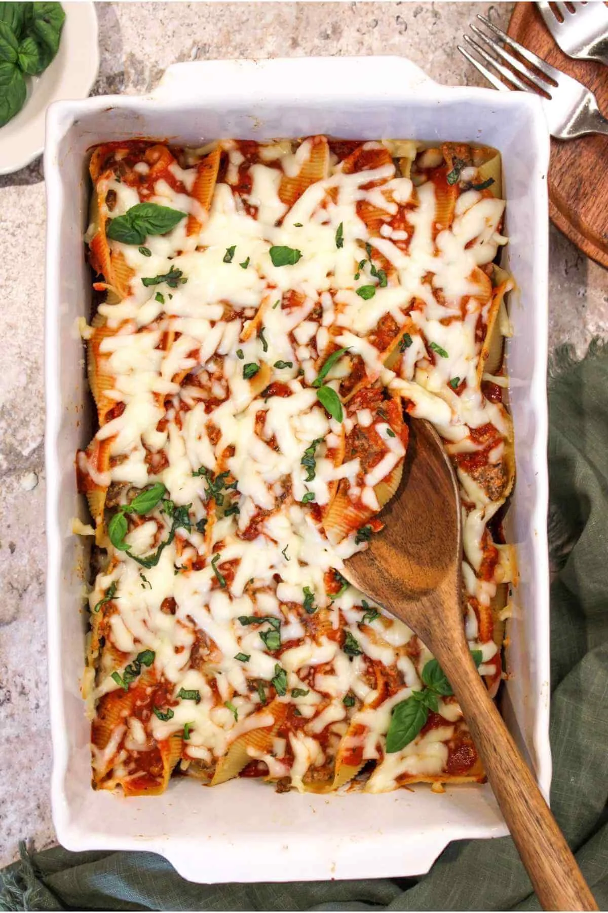 Overhead view of ground beef and ricotta stuffed shells topped with melted mozzarella cheese and fresh basil. Wooden spoon to scoop and a white baking dish show a delicious baked dinner recipe.