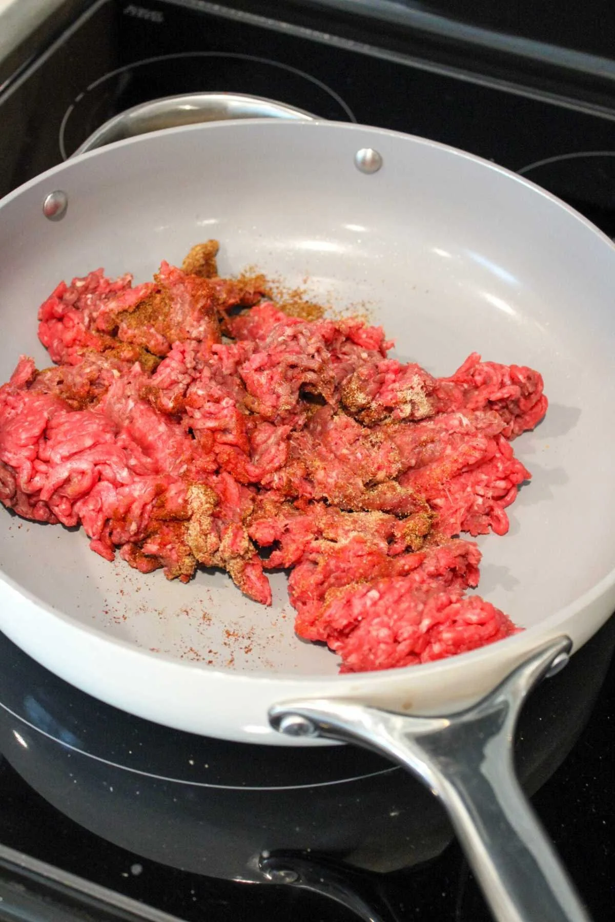 Raw ground beef with seasoning in a white frying pan on a stove, ready to be cooked. The bright red color indicates it's uncooked.