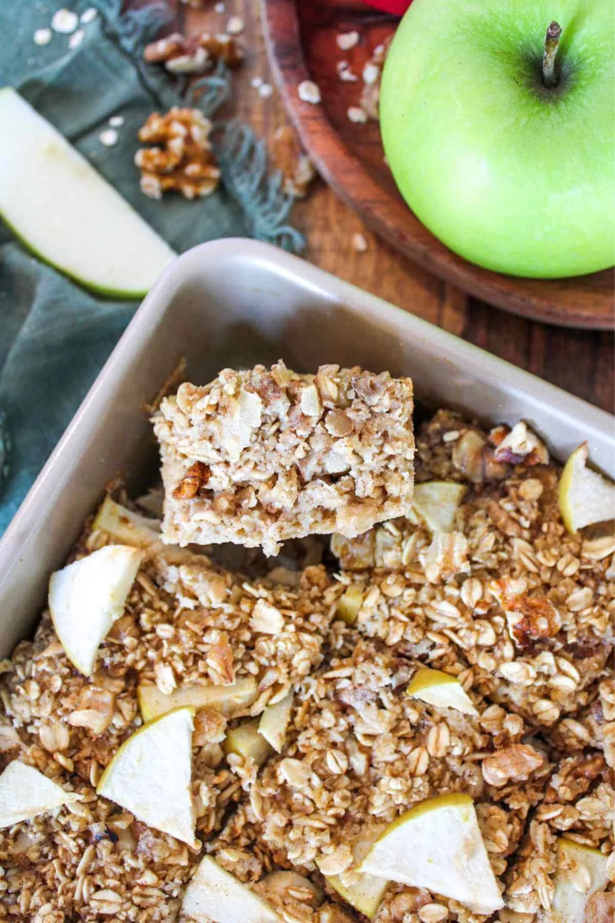 Golden baked apple-oat bars with crunchy granola and apple pieces in a square pan, beside a bright green apple, walnuts, and cinnamon.