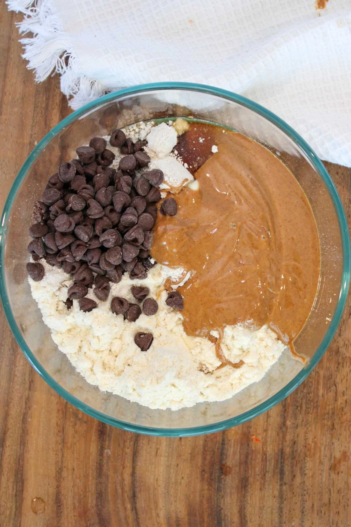 A glass bowl on a wooden surface contains chocolate chips, almond butter, protein powder, and a white cloth in the background. Ingredients are ready to be mixed.