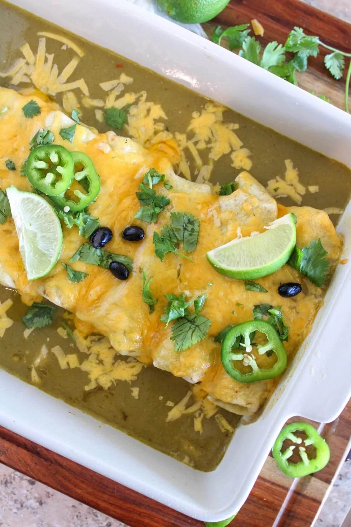 Golden baked chicken enchiladas in green chile sauce, topped with melted cheese, black beans, cilantro, jalapeño slices, and lime wedges.