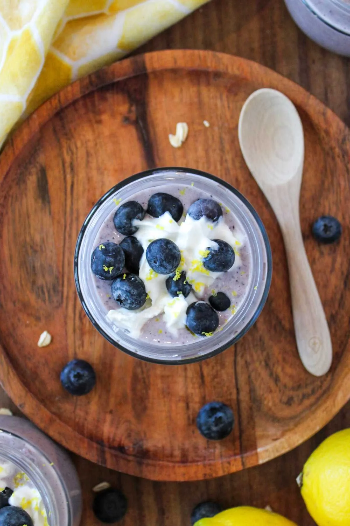 A jar of blueberry overnight oatmeal topped with fresh blueberries and lemon zest on a wooden plate. A wooden spoon and scattered blueberries add a fresh, cozy vibe.