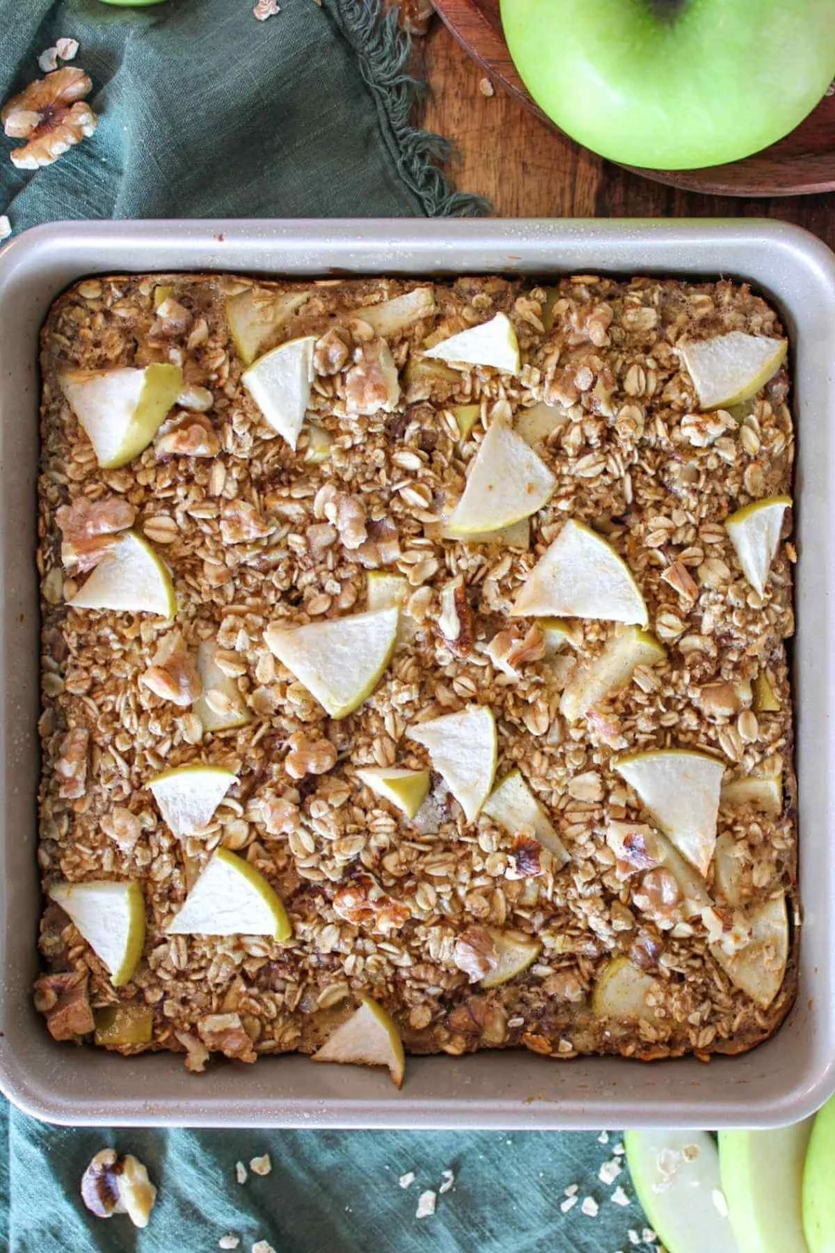 Overhead view of golden baked apple oat bars in a square pan, topped with apple slices, on a rustic wood table with green apples.