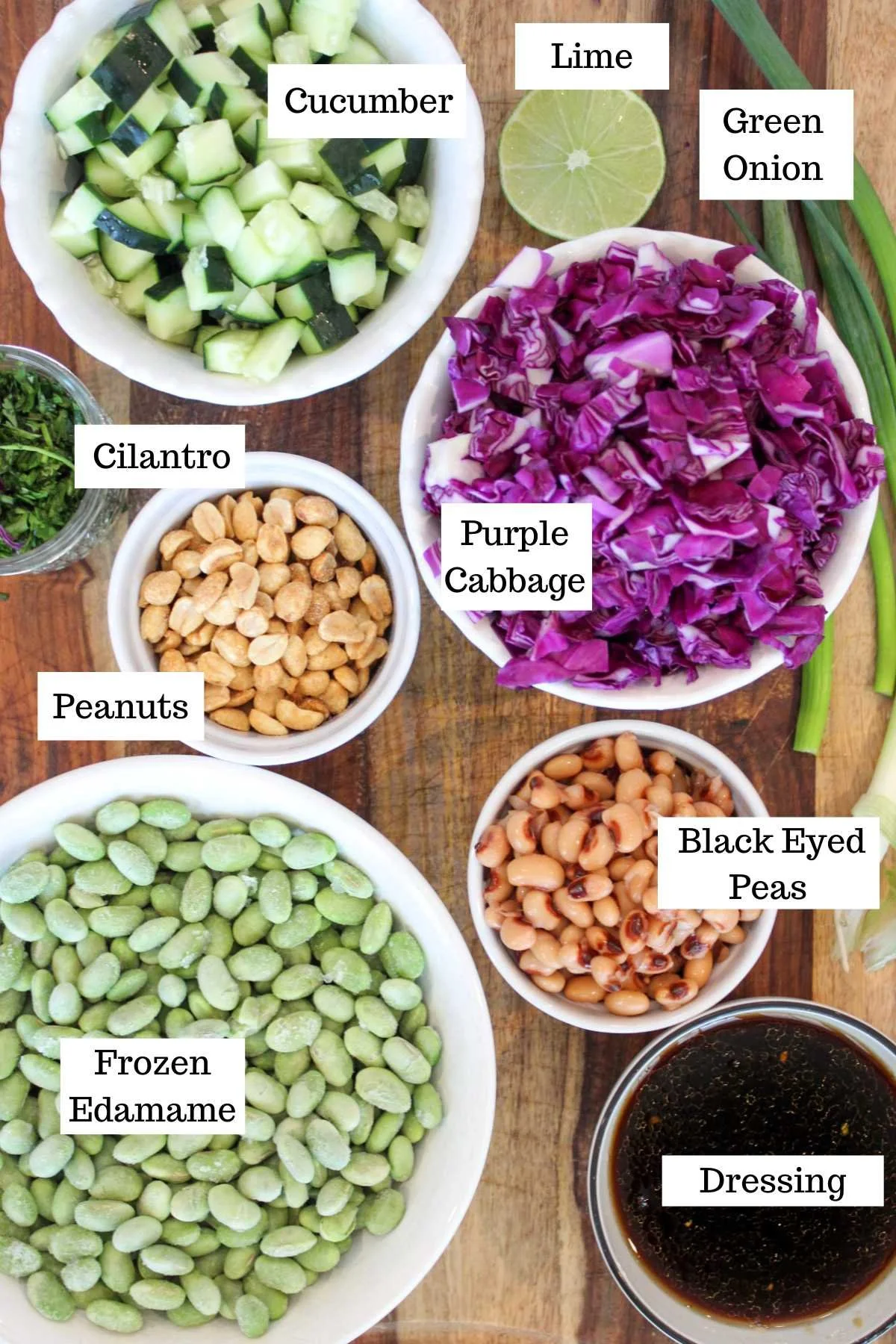 Bowls of cucumber, lime, green onion, cilantro, purple cabbage, peanuts, edamame, black-eyed peas, and dressing on a wooden surface.