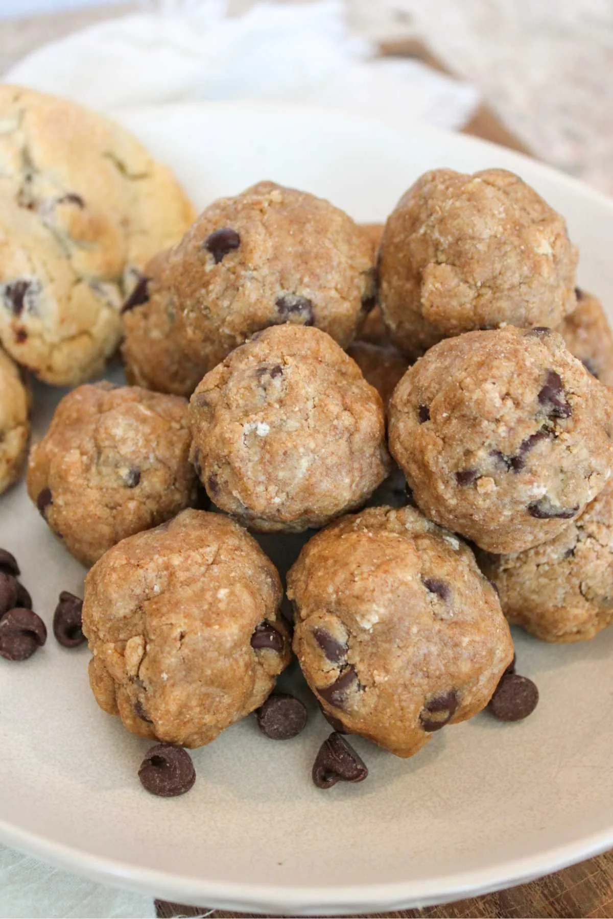 A plate of round, golden-brown chocolate chip cookie protein balls piled together. They are surrounded by scattered chocolate chips, evoking a warm, homemade feel.