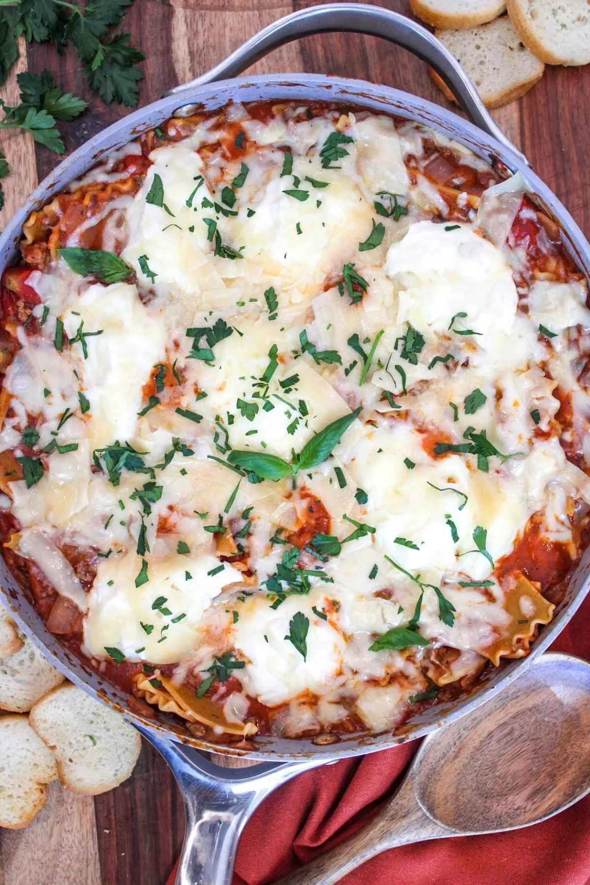 Overhead view of large skillet filled with deconstructed lasagna recipe and topped with melted parmesan cheese and fresh herbs. Side of baguette displayed on wooden backdrop.