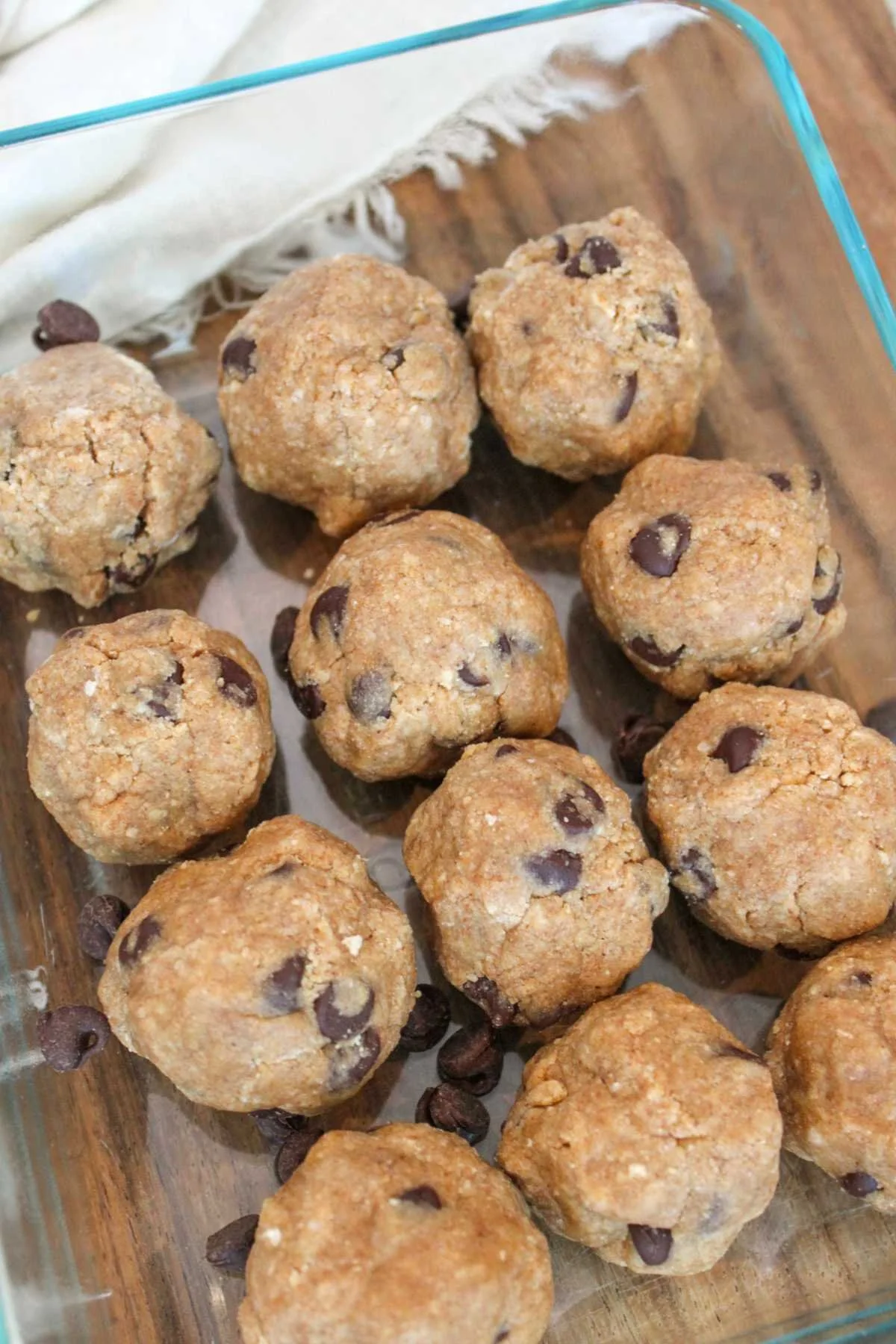 Glass dish with twelve raw chocolate chip protein cookie dough balls. They sit on a wooden table with a white cloth nearby, evoking a cozy feel.