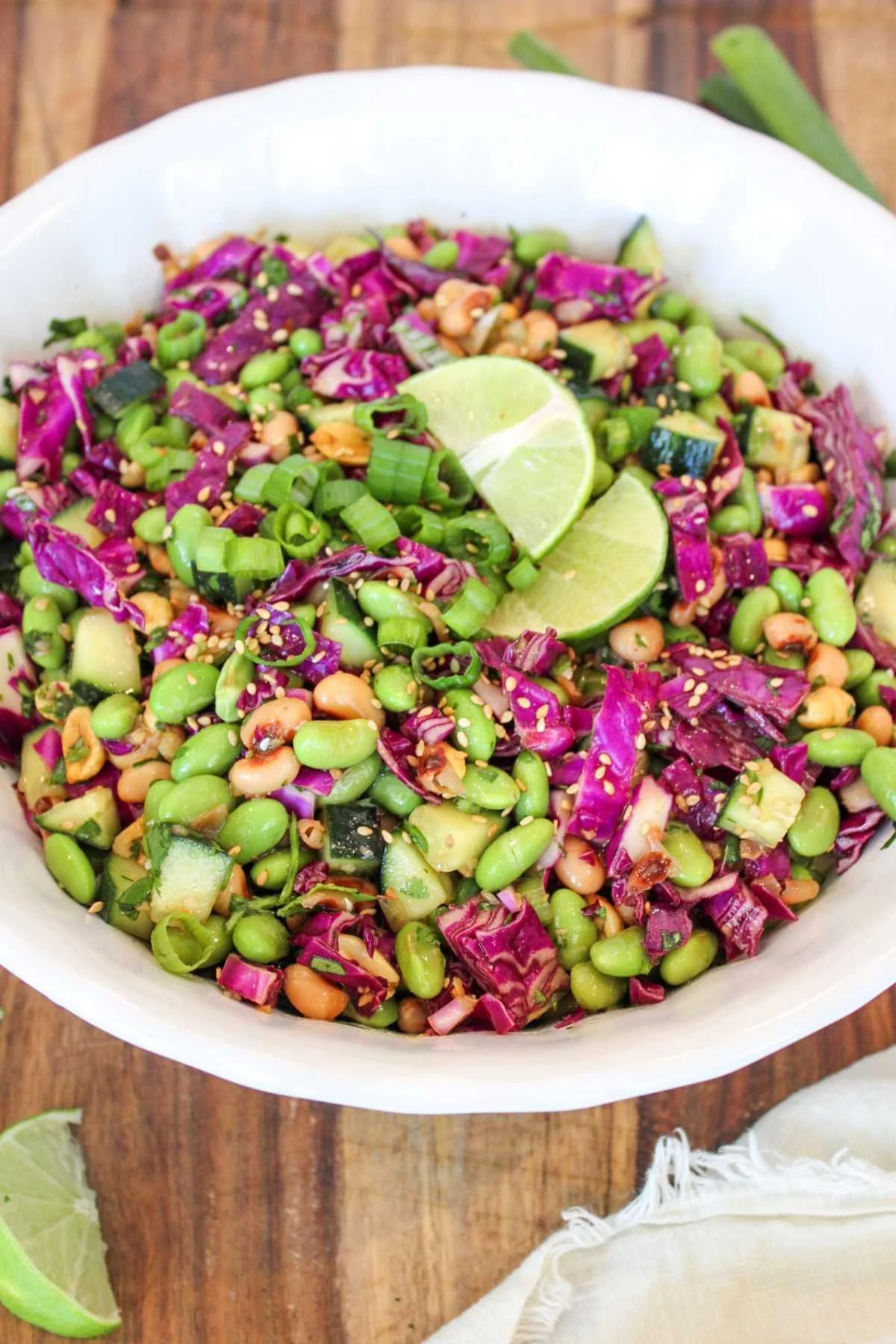 A vibrant salad with purple cabbage, edamame, peanuts, cucumber, and green onions, garnished with lime slices and sesame seeds in a white bowl.