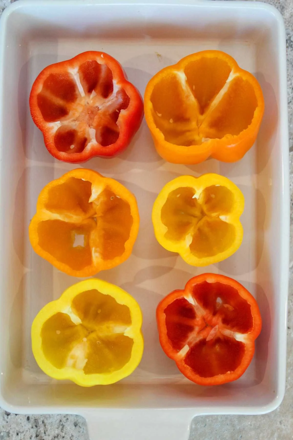 Six halved bell peppers in a white dish: red, orange, and yellow. Bright, fresh tones suggest a lively and healthy cooking atmosphere.
