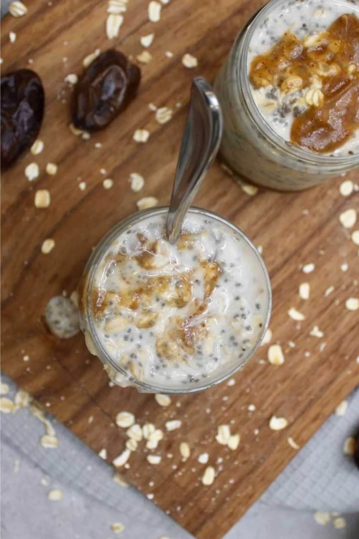 Two jars of overnight oats topped with caramel and chia seeds sit on a wooden board. Dates and scattered oats add a rustic touch to the scene.