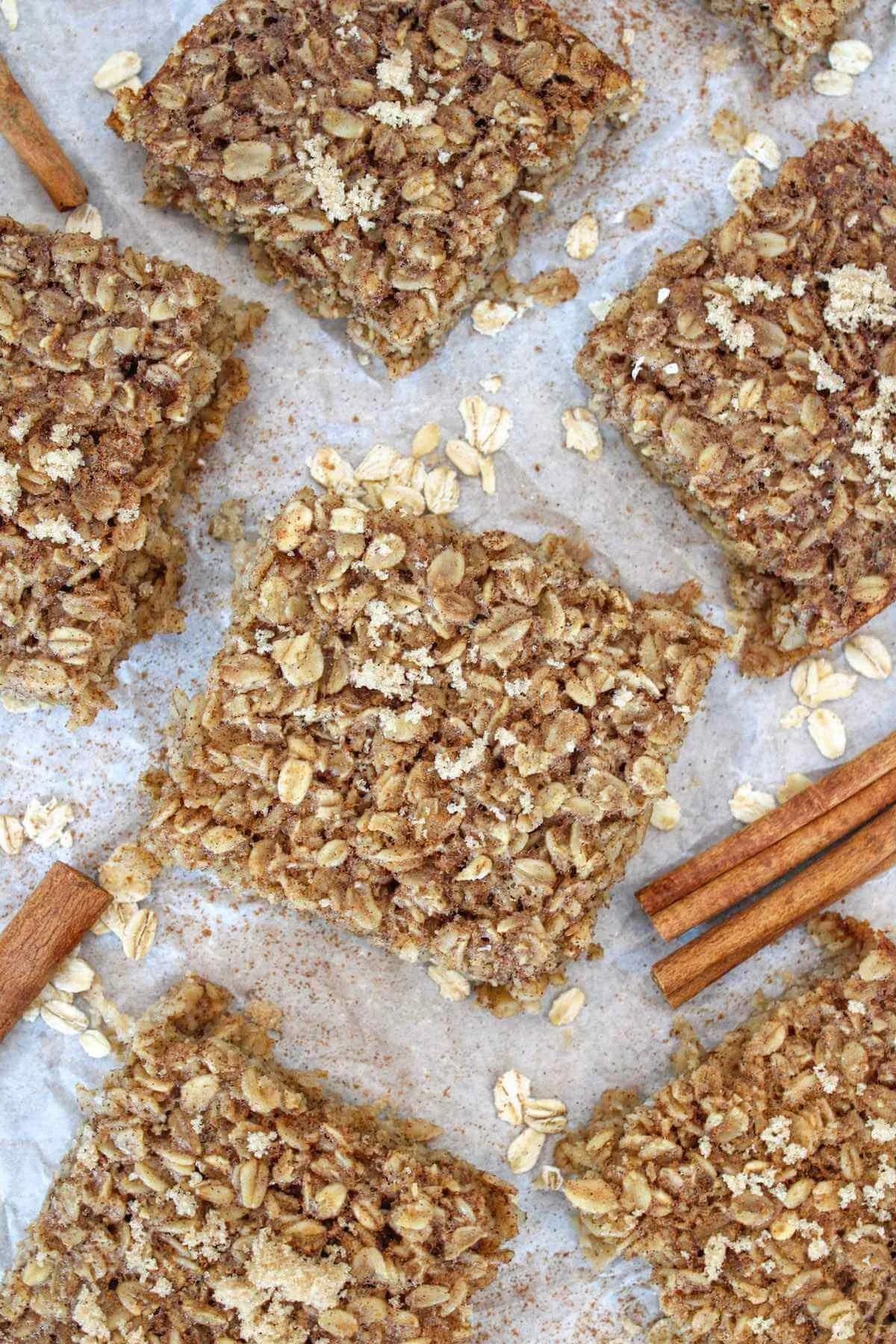 Overhead view of rustic oat granola bars on parchment, sprinkled with oats and cinnamon sticks, creating a warm, homemade baking scene.