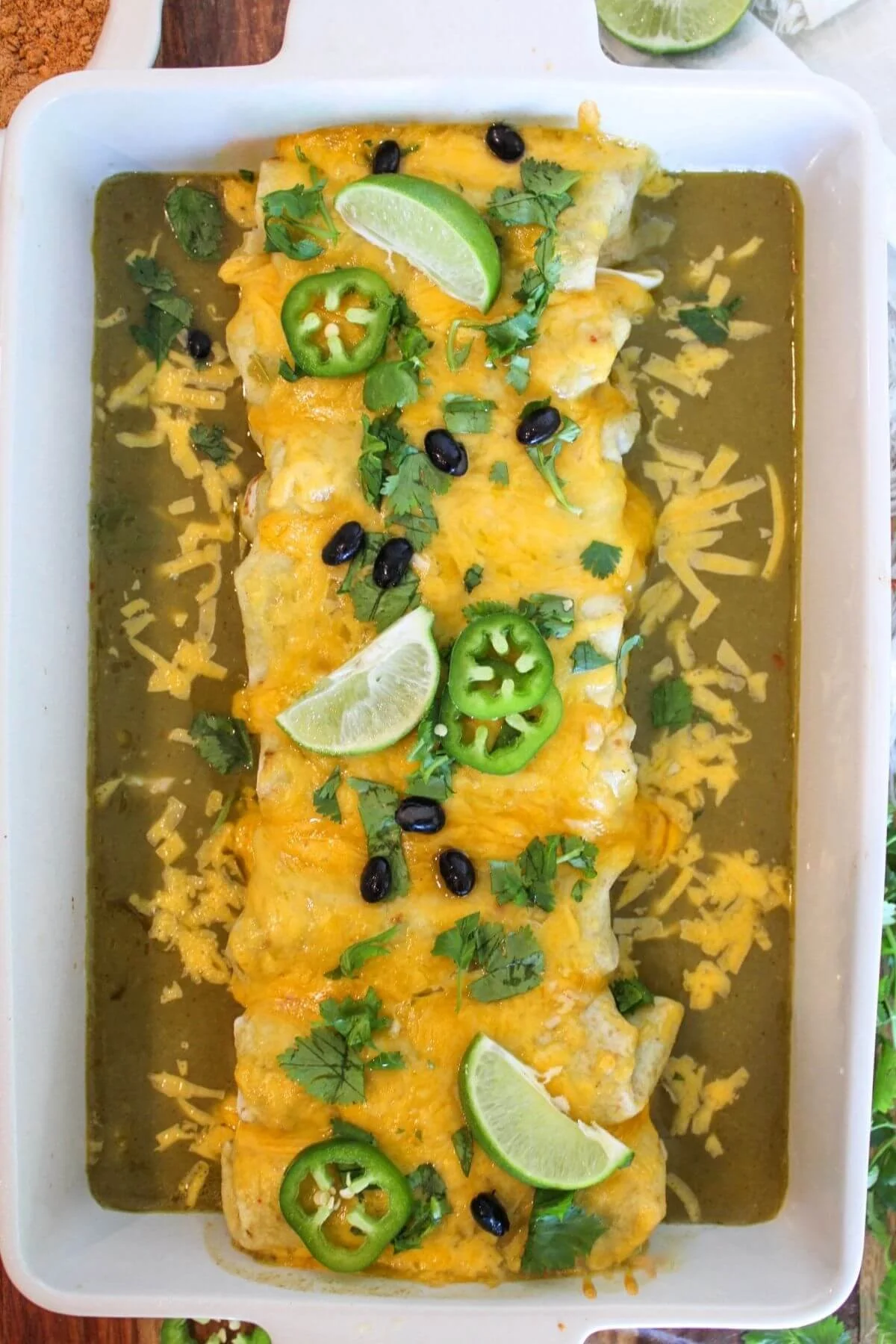 Cheesy chicken and black bean enchiladas in green sauce, topped with black beans, jalapeño slices, cilantro, and lime wedges in a white baking dish.