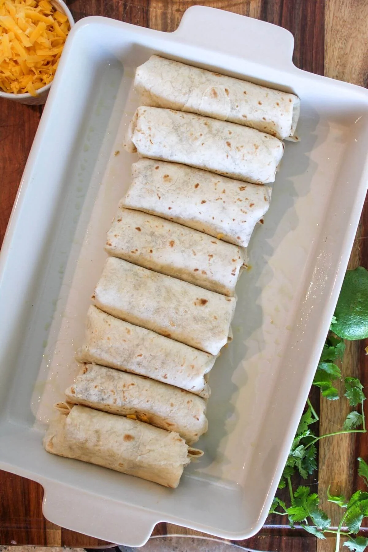 Overhead view of eight golden-brown rolled chicken enchiladas in flour tortillas arranged in a white baking dish, with shredded cheese and fresh cilantro nearby.