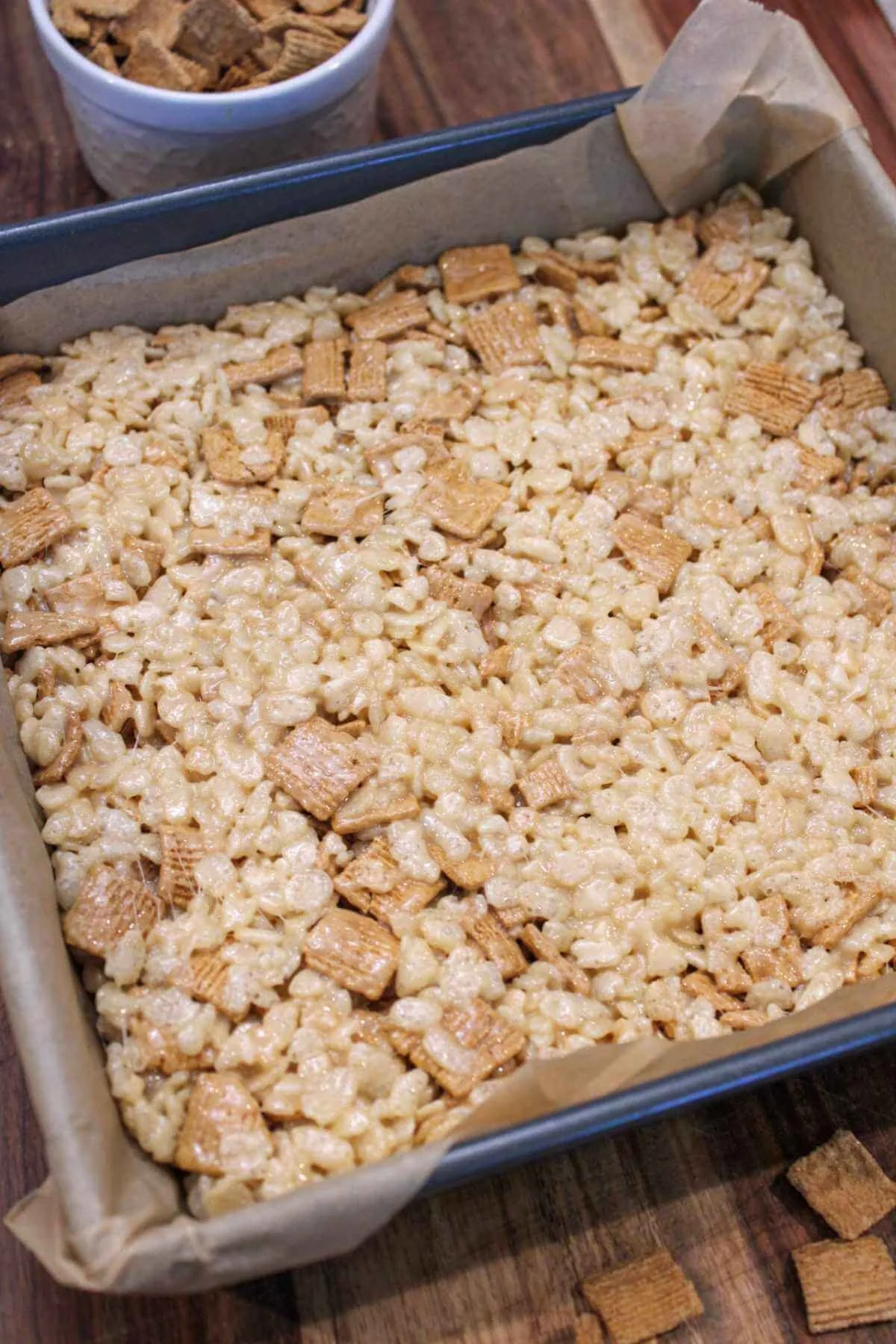 8x8 metal pan is lined with parchment paper and filled with brown butter Rice Krispie treats with Golden Grahams and served on a wooden cutting board with a side cup of golden graham cereal.