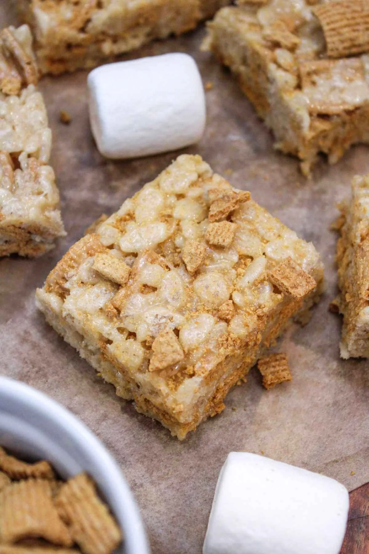 brown butter rice krispie treat with golden graham cereal is served on parchment paper with surrounding bars and marshmallows.