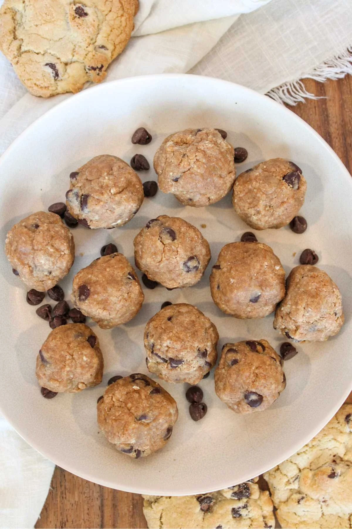 A white plate holds several round chocolate chip cookie dough protein balls. Surrounding the plate are scattered chocolate chips and a baked cookie, set on a wooden table with linen cloths, evoking a cozy, homemade feel.