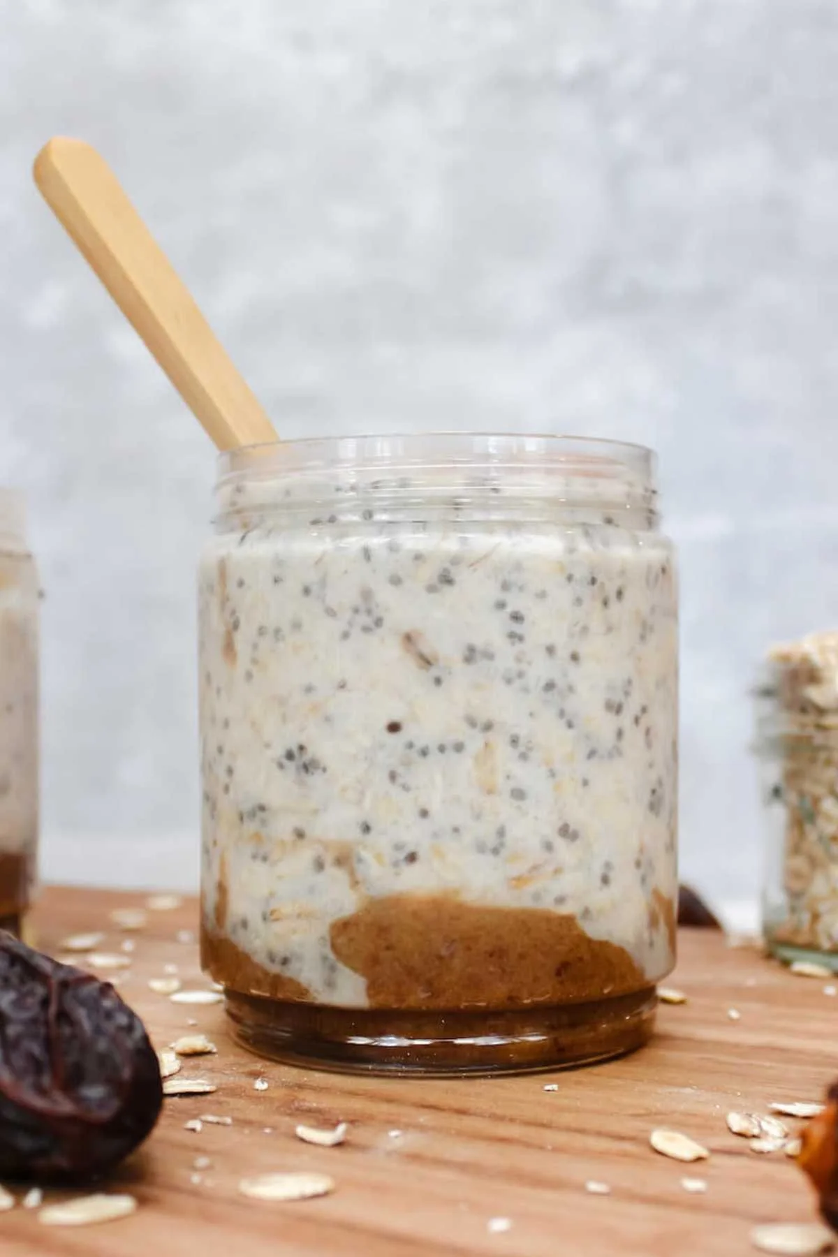 A jar of overnight oats with chia seeds, layered over a smooth nut butter. A wooden spoon is inserted. Scattered oats and a date rest on a wooden surface.