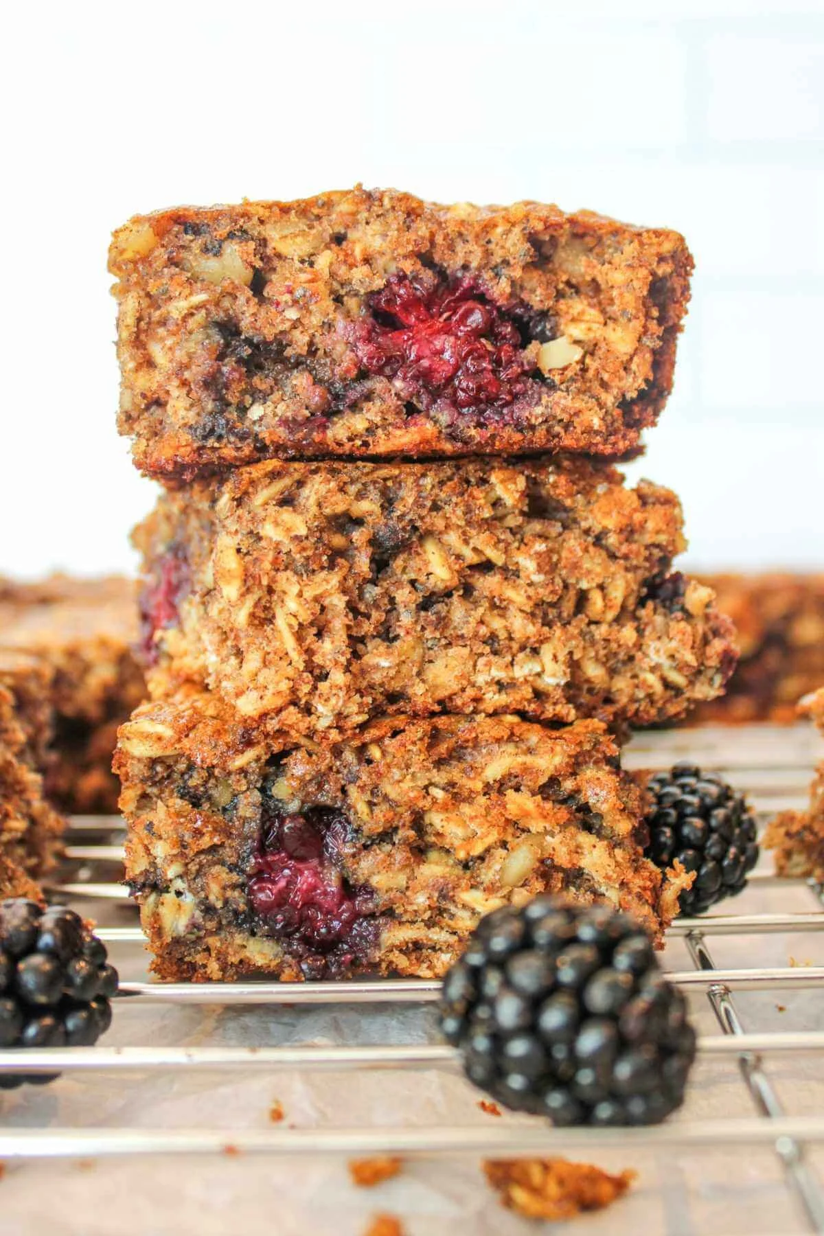 Stack of three golden-brown blackberry oatmeal bars on a cooling rack, with juicy berry centers and fresh blackberries in the foreground.