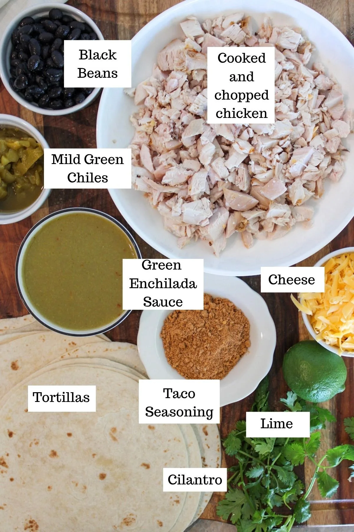 Overhead layout of chicken enchilada ingredients: chopped chicken, black beans, green chiles, enchilada sauce, cheese, tortillas, taco seasoning, lime, cilantro.
