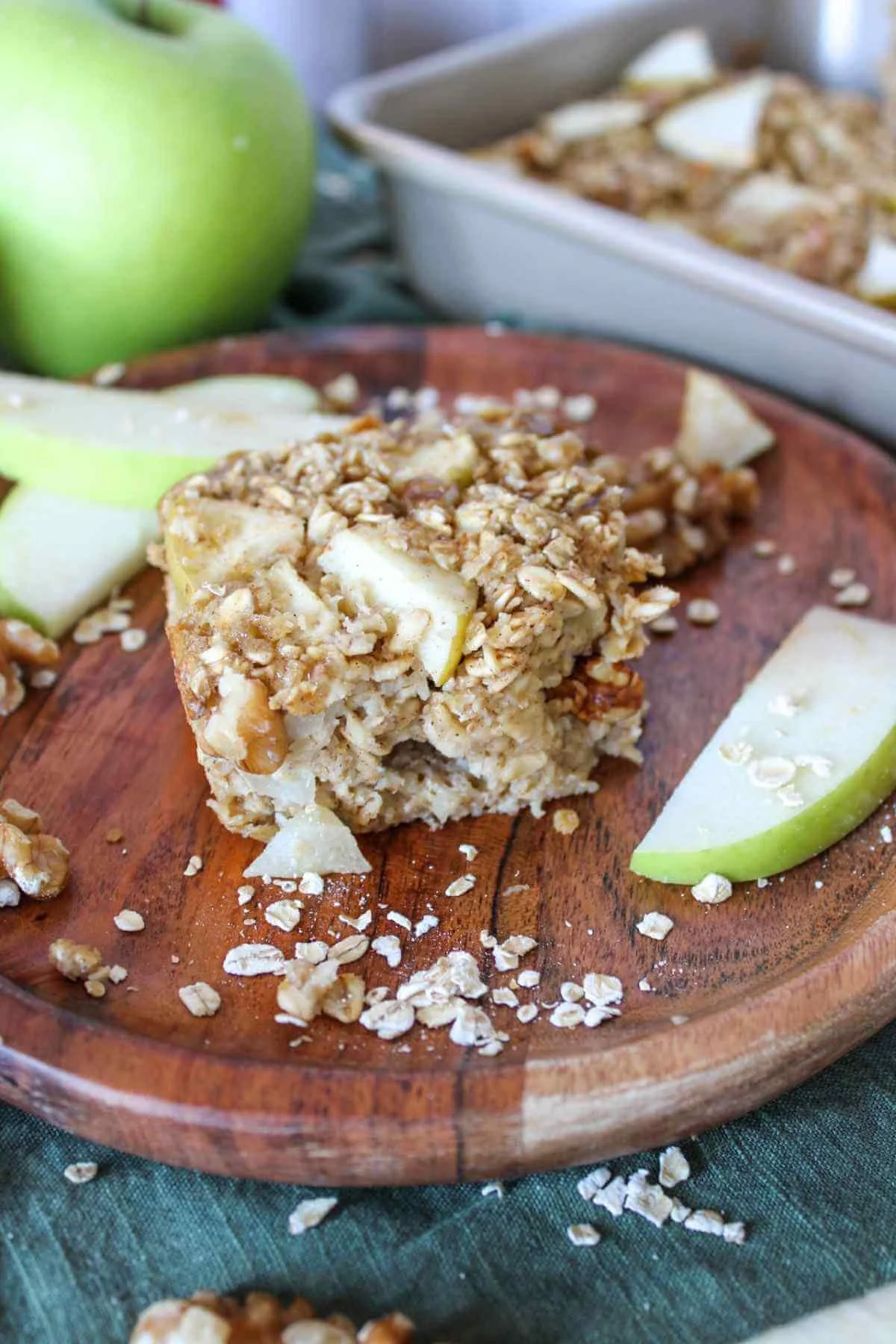 Close-up of a golden apple oatmeal bake on a wooden plate, with green apple slices, scattered oats, and a baking dish in the background.