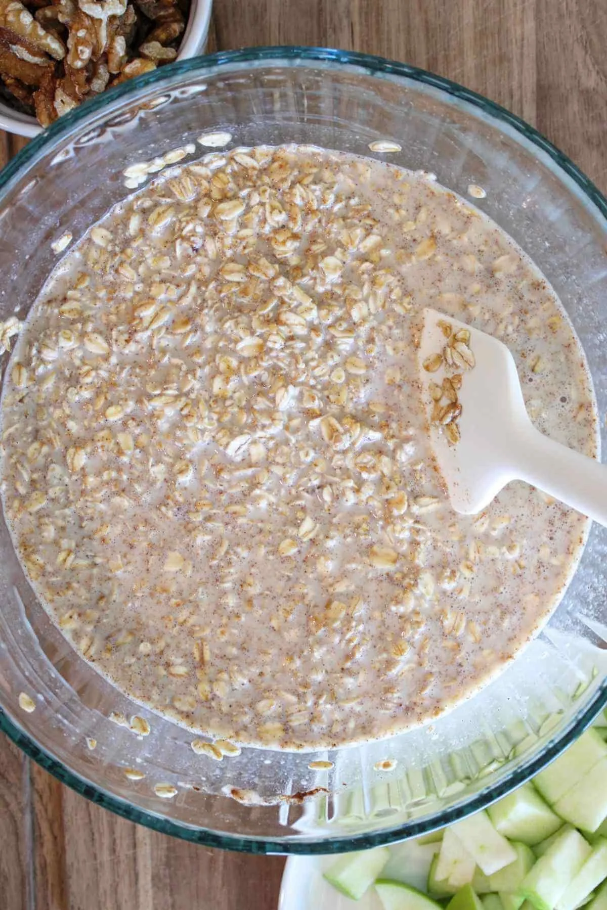 Overhead view of creamy oat mixture in a glass bowl, with a white spatula, walnuts above, and chopped green apples below.