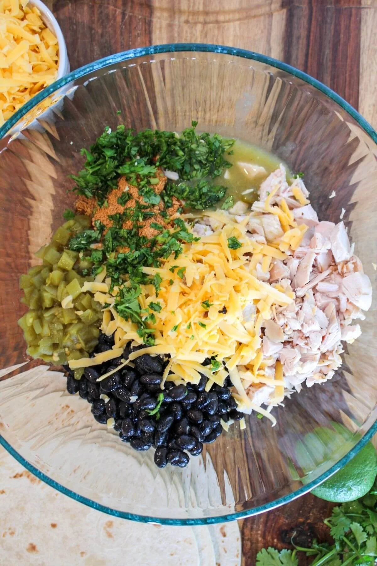 Overhead view of a glass bowl with shredded chicken, cheddar cheese, black beans, green chiles, herbs, and spices—a colorful, appetizing meal.